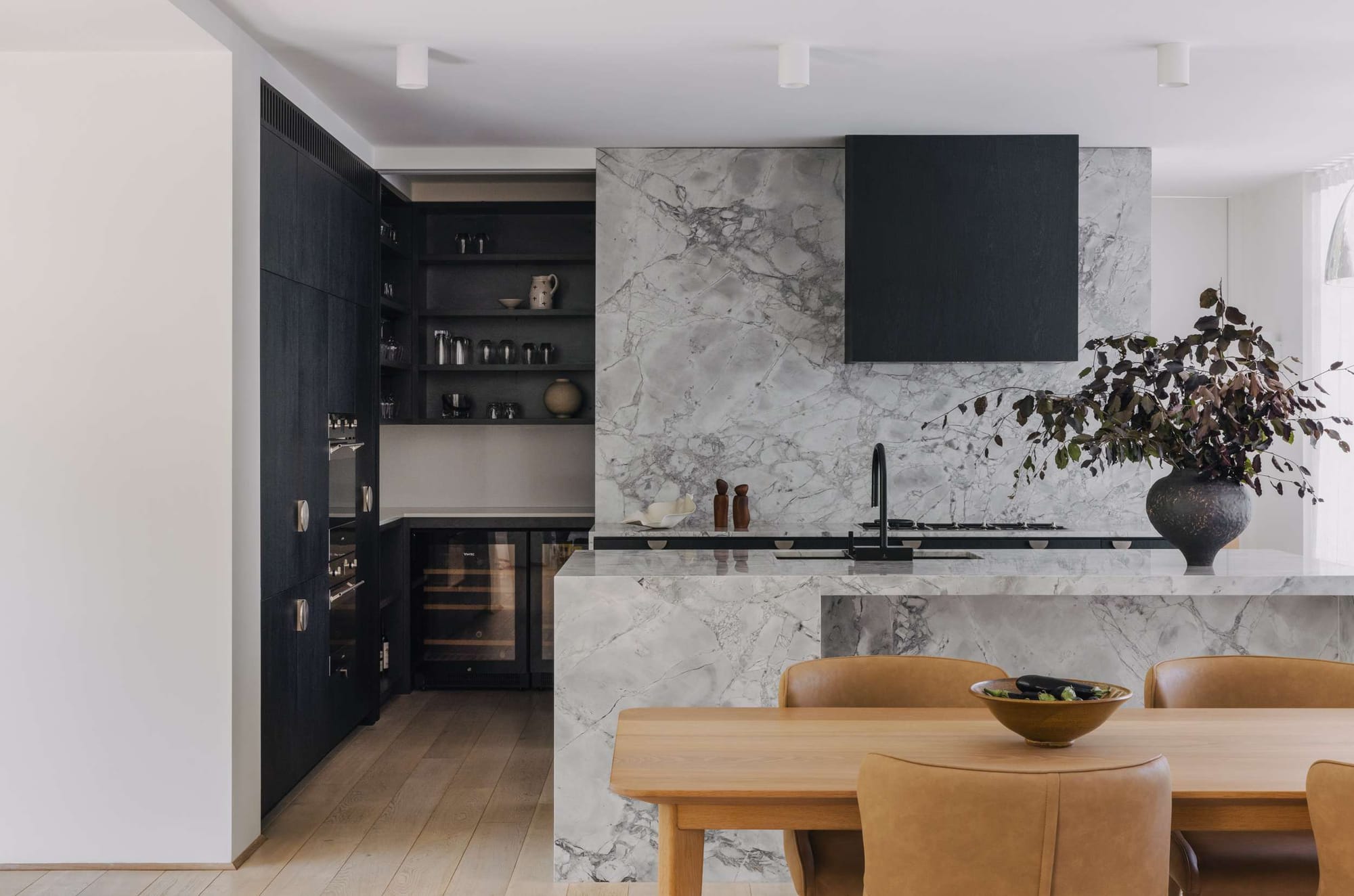 Monochrome kitchen with dramatic veined stone splashback and island, dark cabinetry and a timber dining table in the foreground.