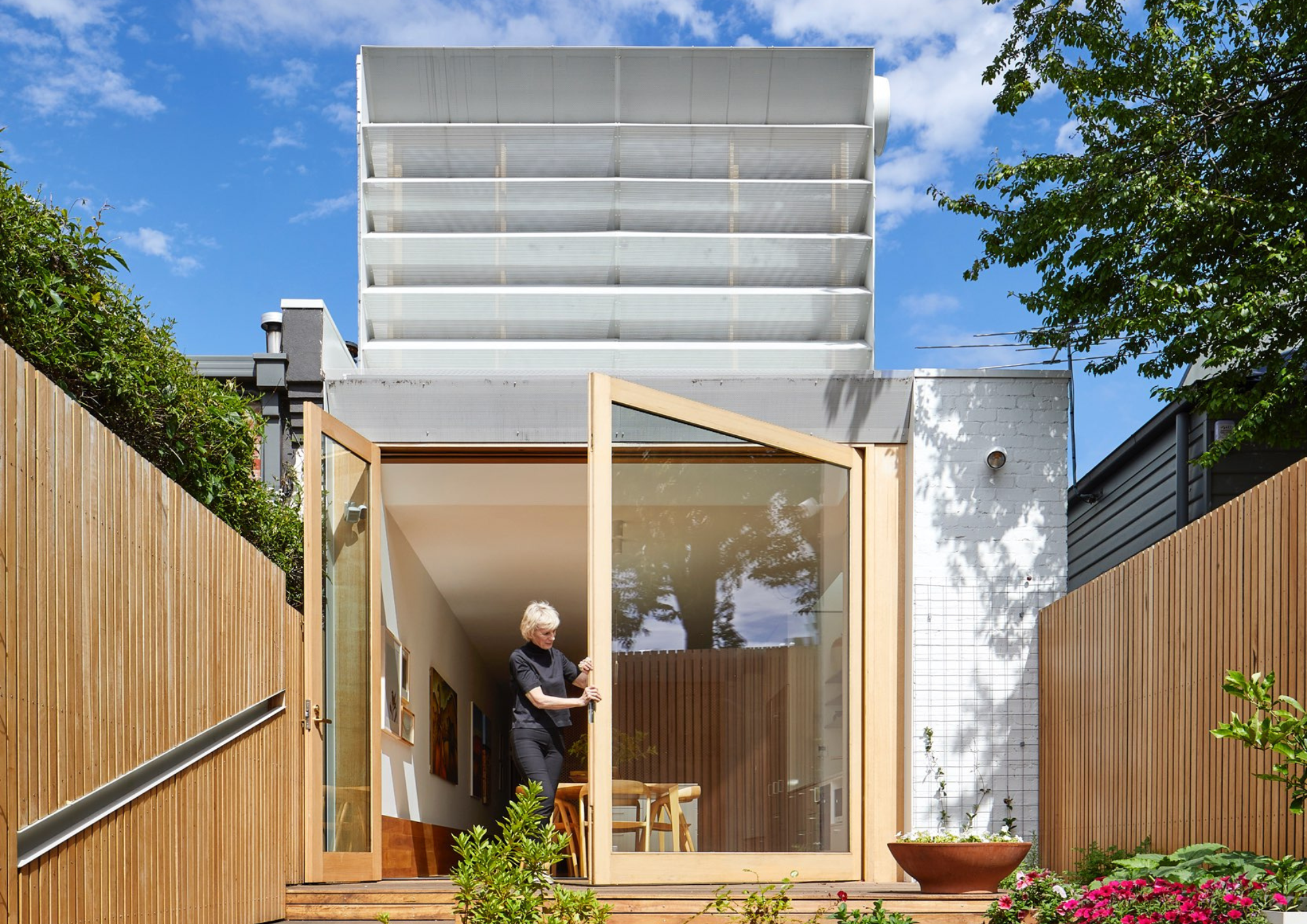 Rear exterior of a contemporary home with large timber-framed glass doors opening onto a garden, topped by a white louvred shading structure.