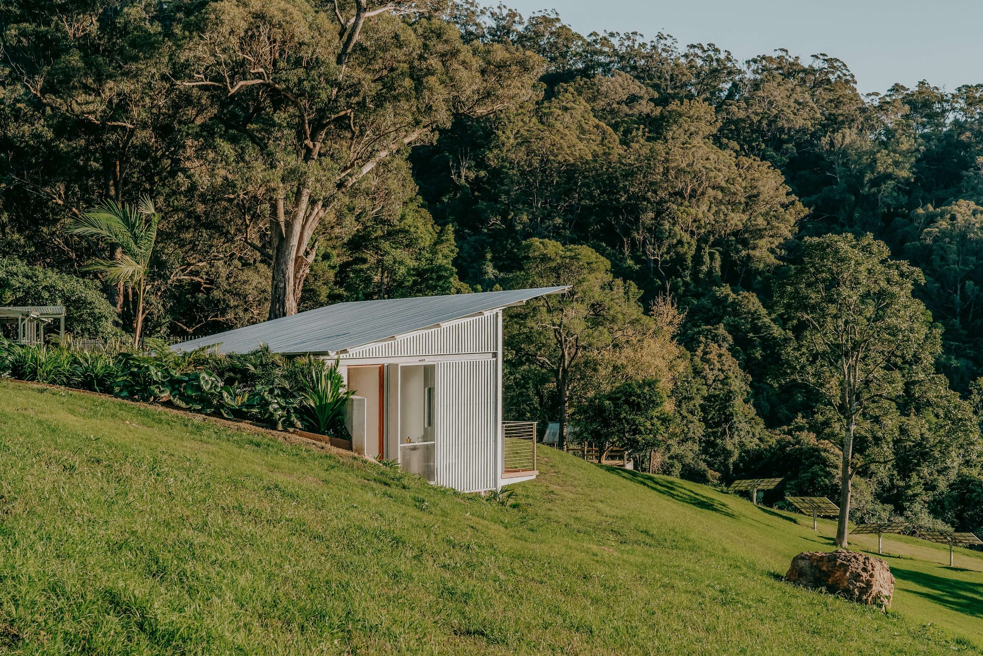Small corrugated metal pool house and sauna by RaddatzKueber positioned on a grassy slope surrounded by bushland.