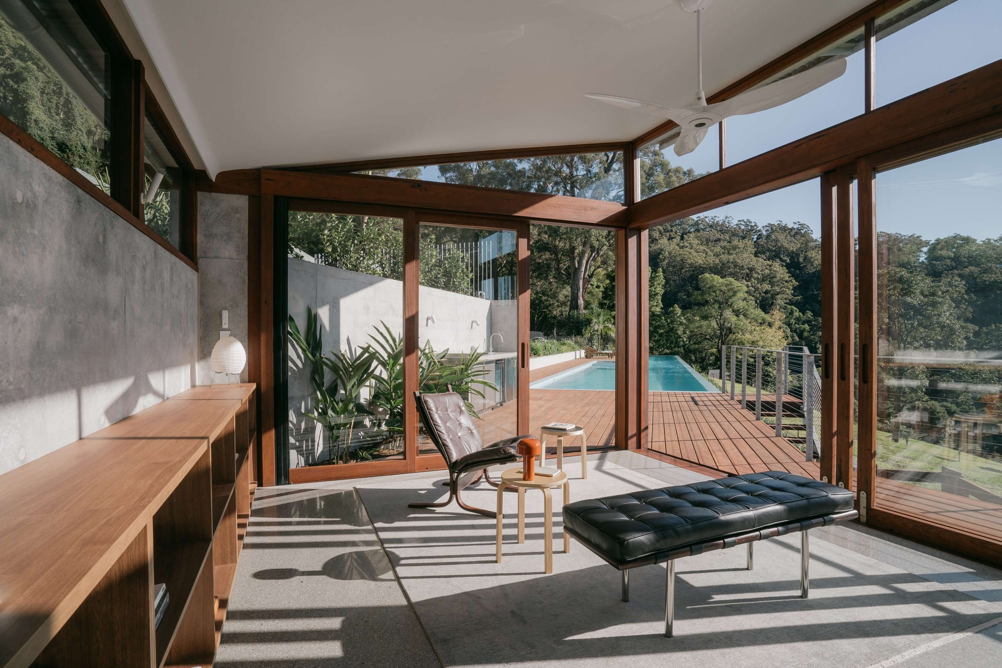 Open sided pavilion style room with timber framing and full height glazing, leather and canvas lounge furniture, opening directly onto a timber deck and elevated lap pool surrounded by bushland.