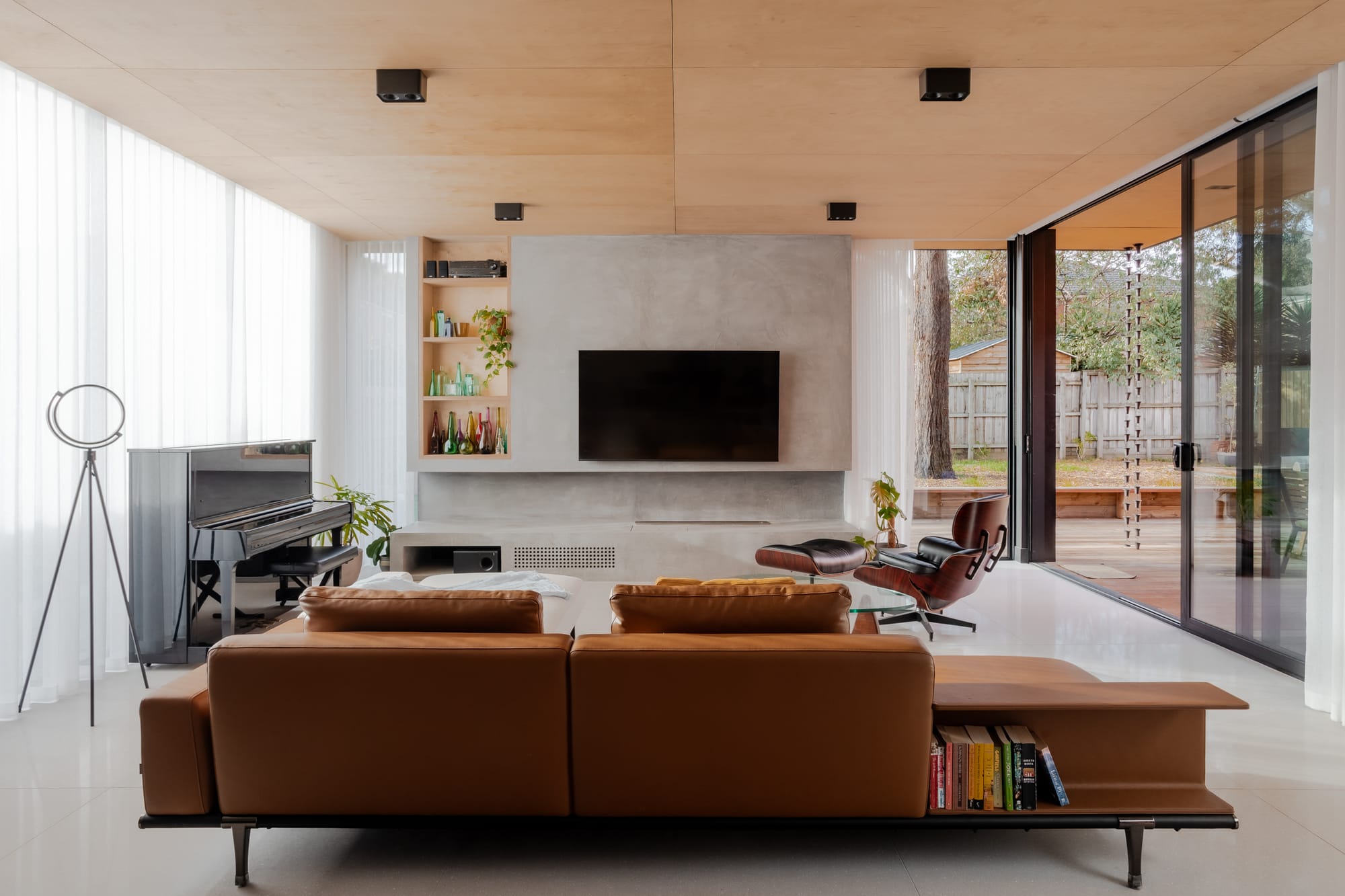 Living room with a plywood ceiling, sheer curtains, tan leather sofa and Eames lounge chair, opening onto a timber deck through black framed sliding doors.