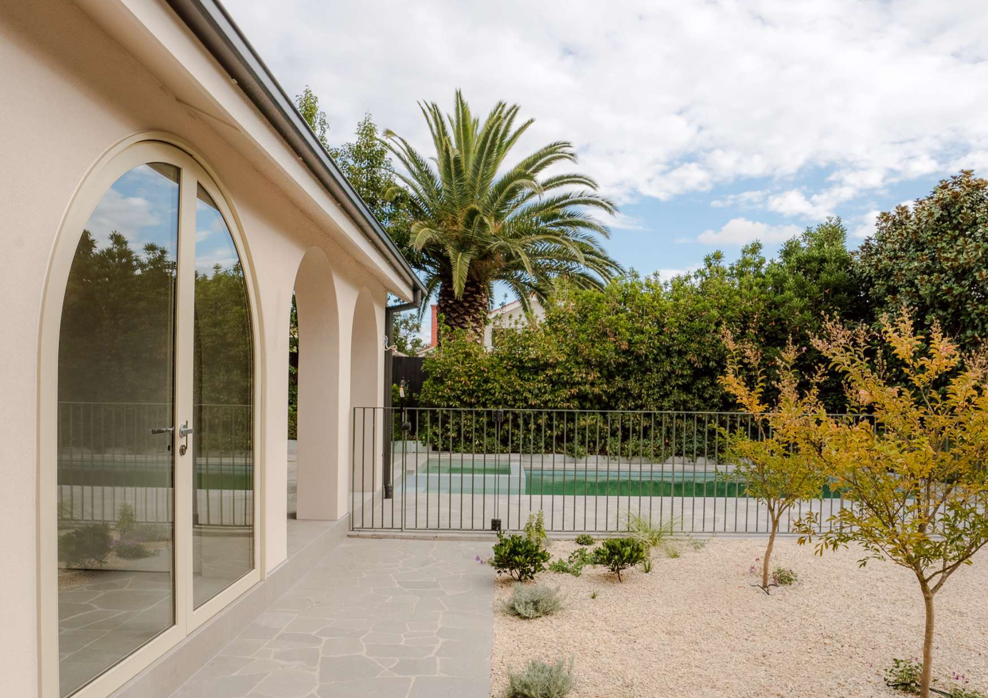 Mediterranean-style courtyard with arched glass doors, pale stone paving, a swimming pool, palm tree and landscaped garden beds.