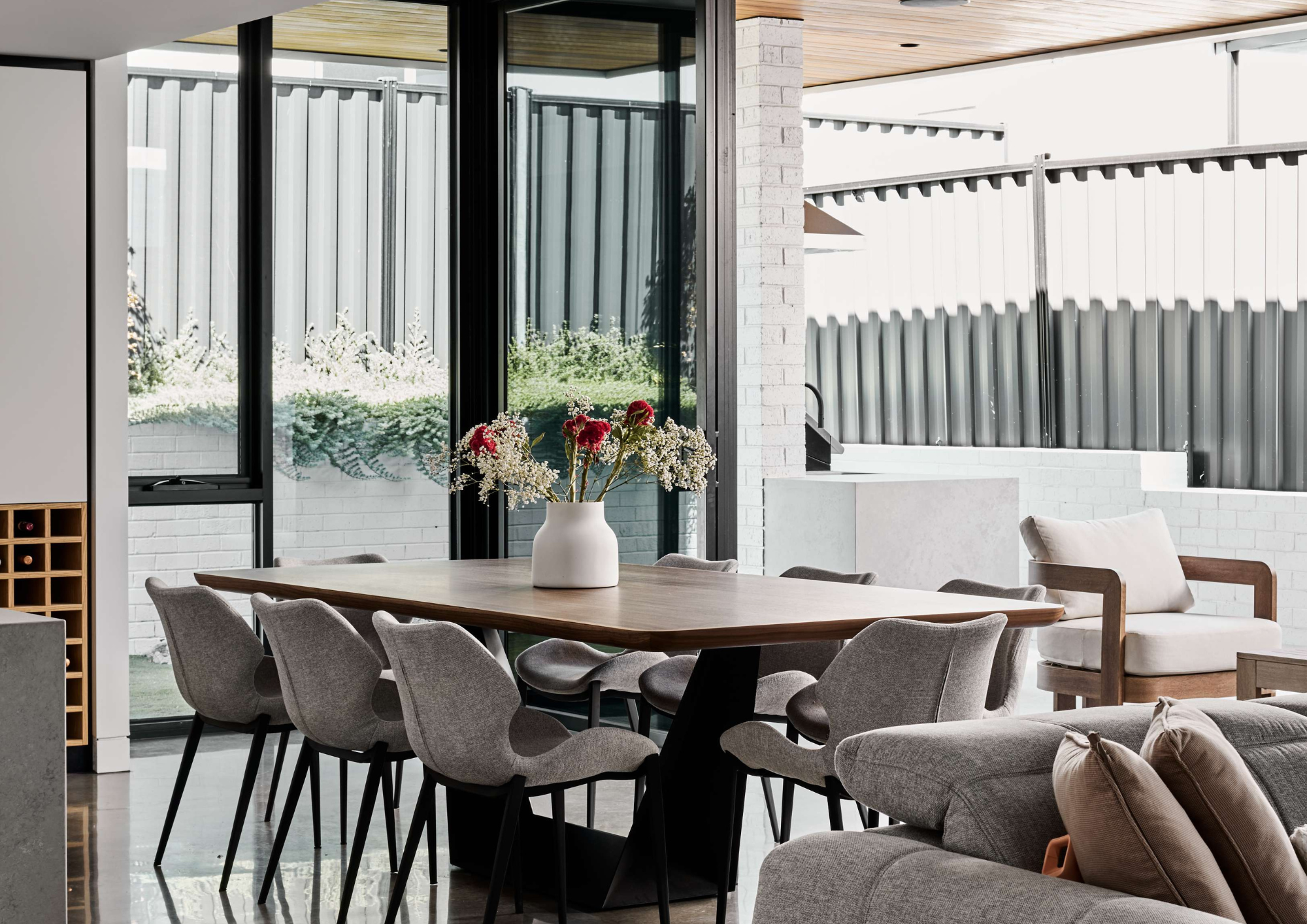 Contemporary dining and living area with a timber dining table, grey upholstered chairs and large black-framed windows overlooking an outdoor courtyard.