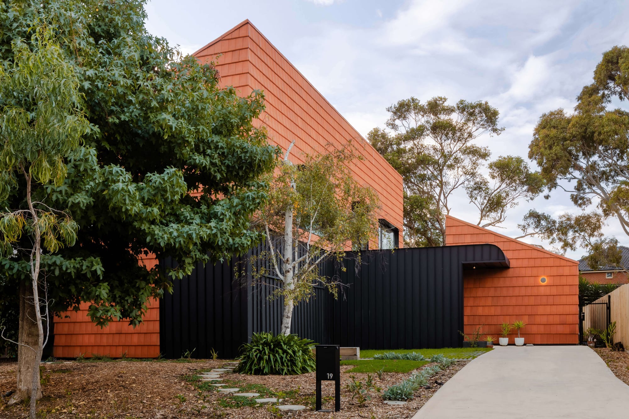 Terracotta tiled house with black cladding and a pitched roof, set among gum trees with a landscaped native garden and concrete pathway.