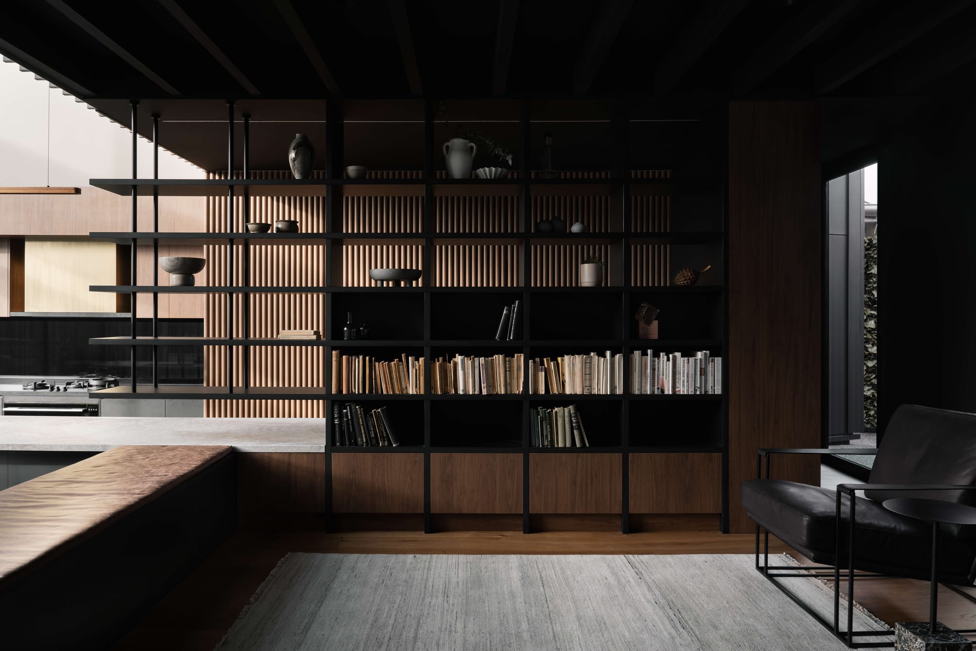 Dark, moody living room with black steel and timber shelving, book-lined shelves and a glimpse of the kitchen lit by an angled glazed roof beyond, Malvern residence