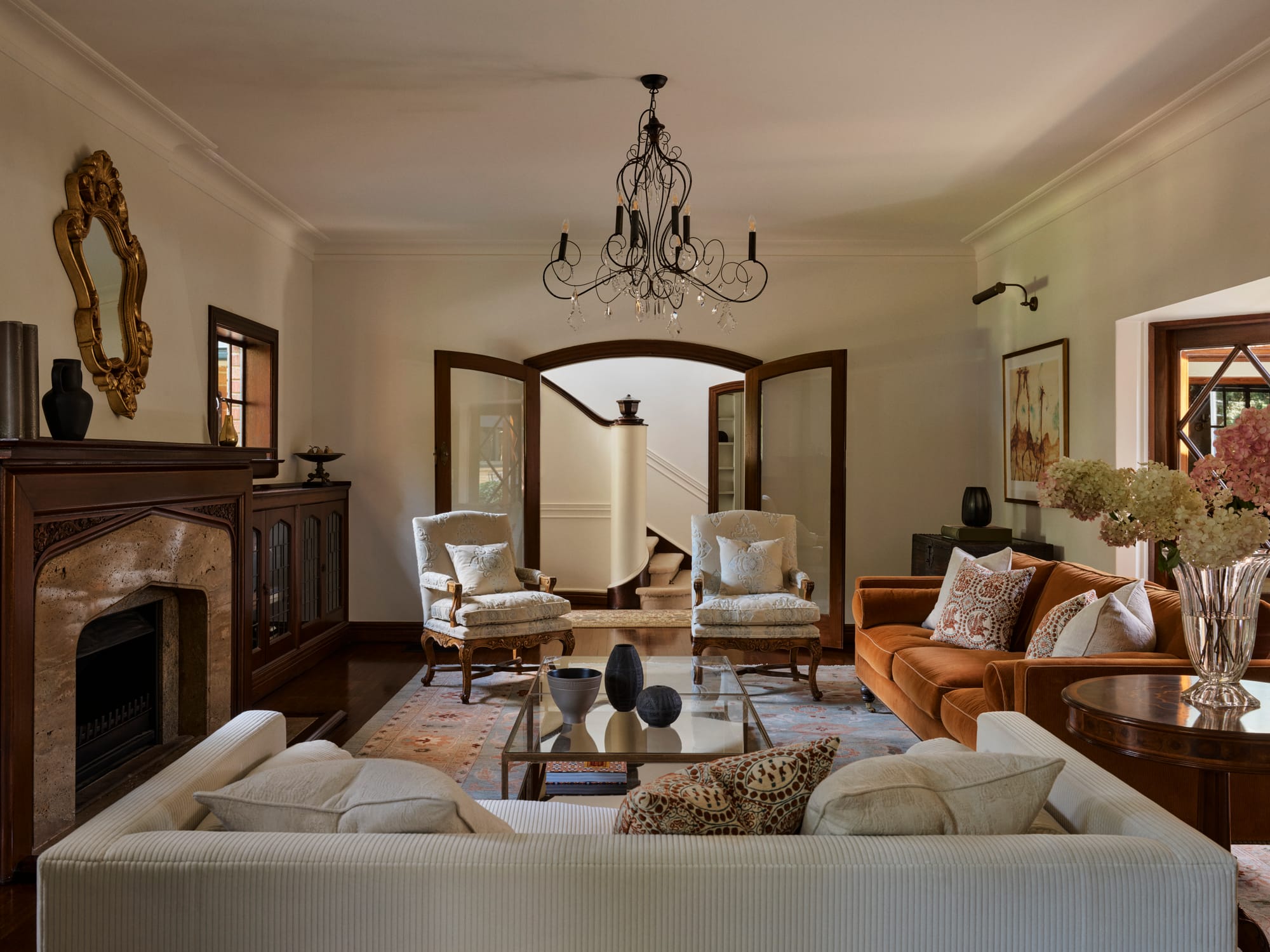 Traditional living room with a carved timber fireplace, ornate gold mirror, chandelier, patterned rug and a rust velvet sofa framed by cream seating.