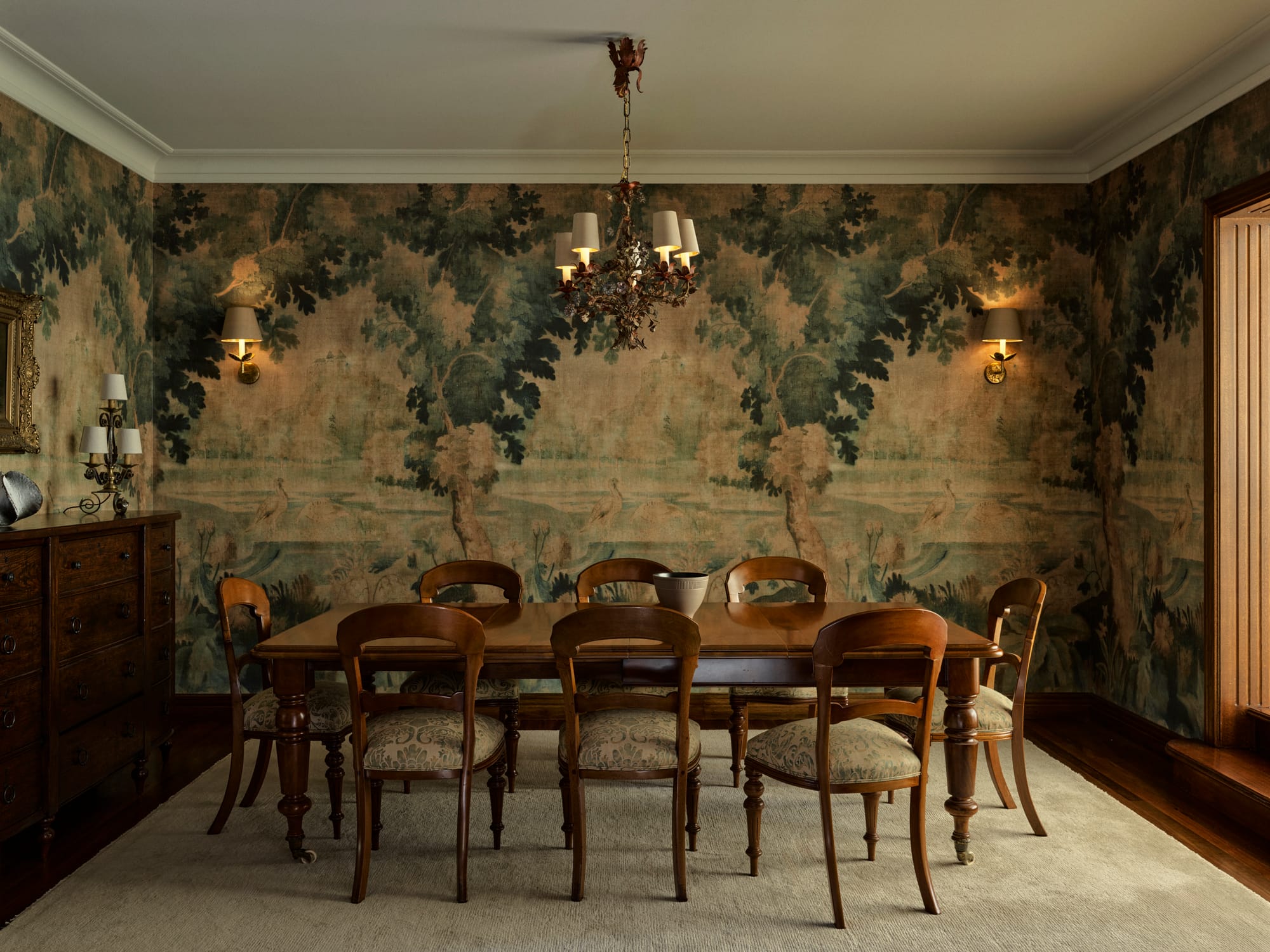 Formal dining room with antique timber furniture, botanical mural-style wallpaper, wall sconces and a decorative chandelier creating a richly layered interior.