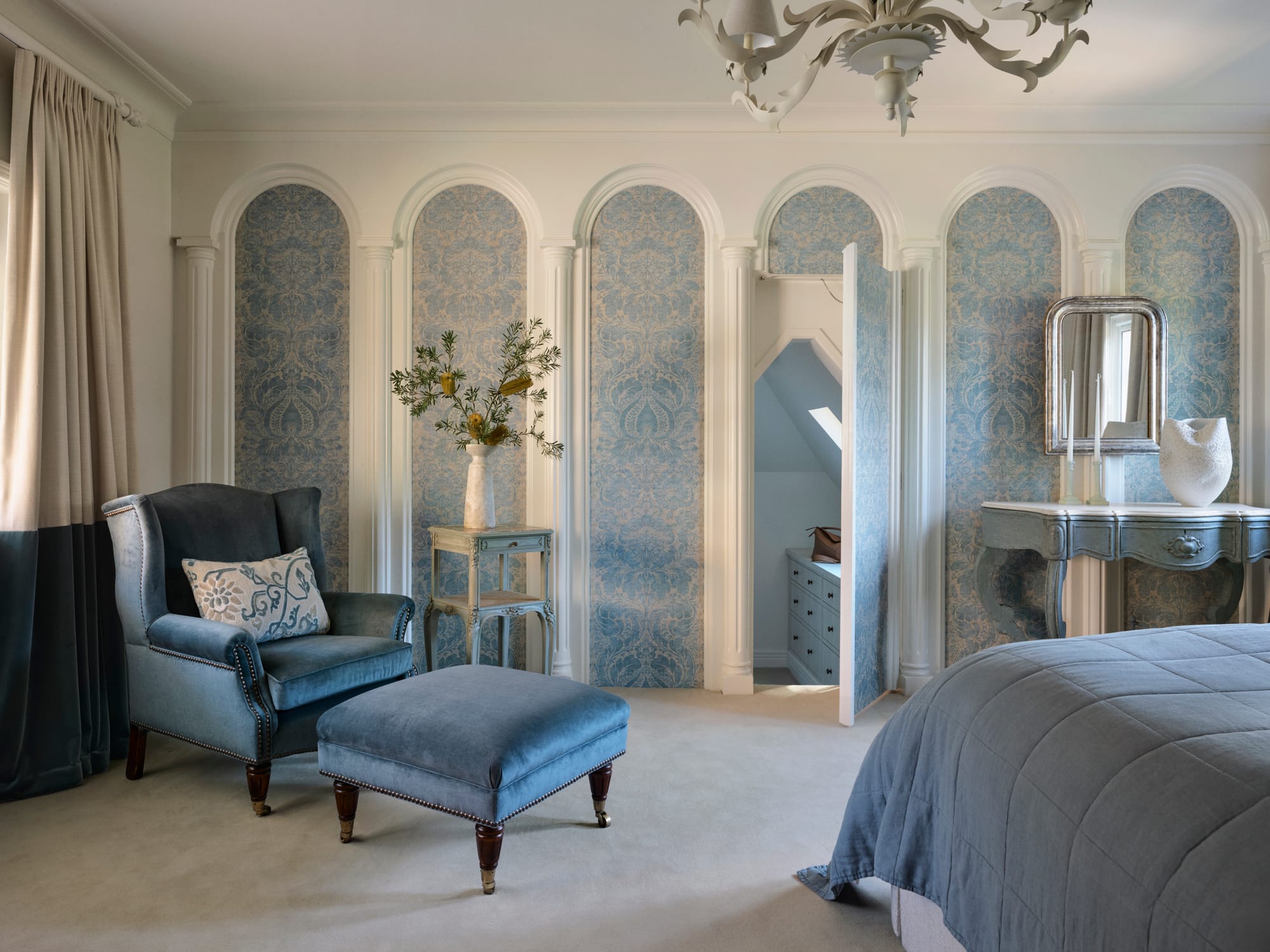 Soft blue bedroom with arched wall panelling, patterned wallpaper, velvet armchair and ottoman, antique-style console and a concealed doorway.