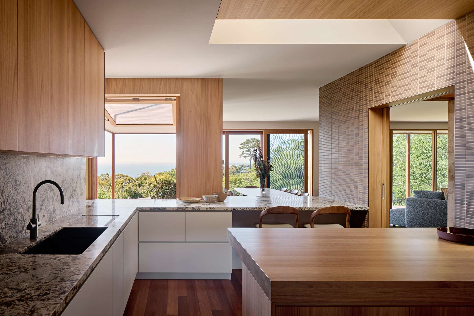 Contemporary kitchen with timber cabinetry, stone benchtops and a large timber island, framed by windows overlooking a green landscape.