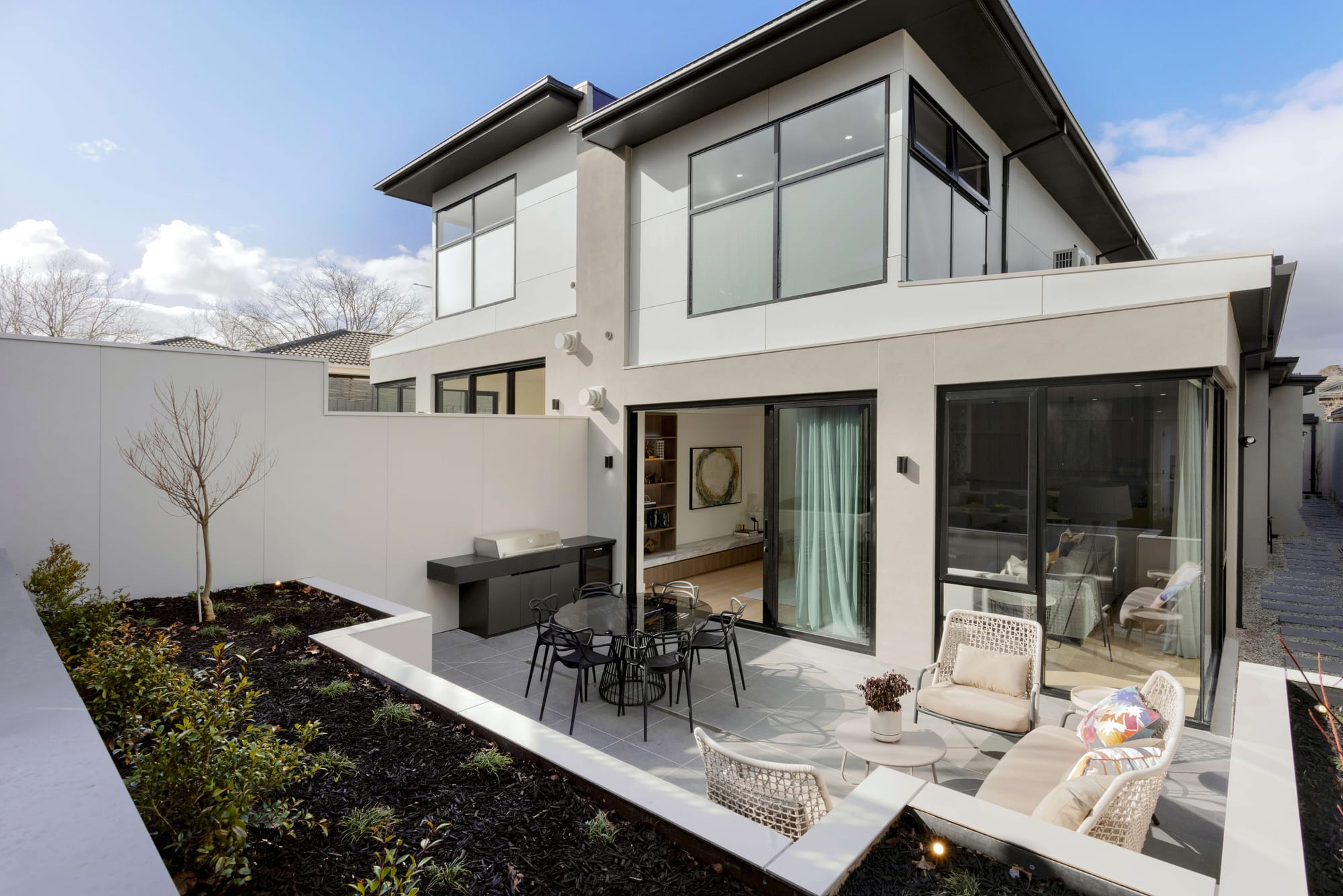 Contemporary two storey home with a sunken courtyard garden, alfresco dining and lounge setting, and a built in outdoor kitchen opening onto a paved terrace framed by dark cladding and large glazed doors.