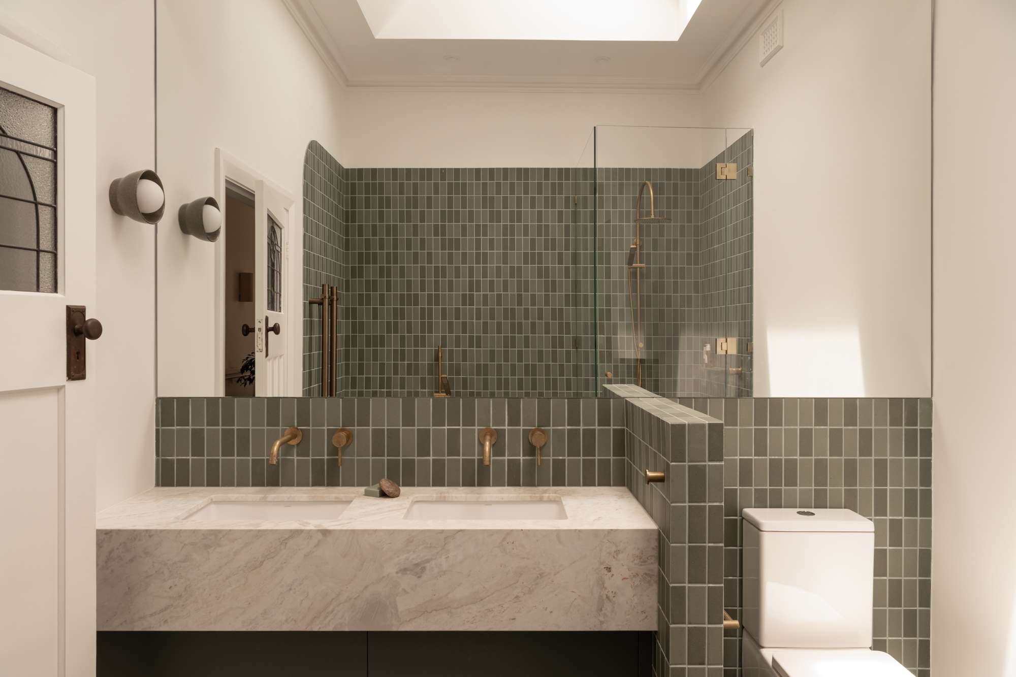 A bathroom with sage green square tiles, a curved tiled shower nook with brass tapware, and a marble double vanity beneath a large mirror lit by a skylight.