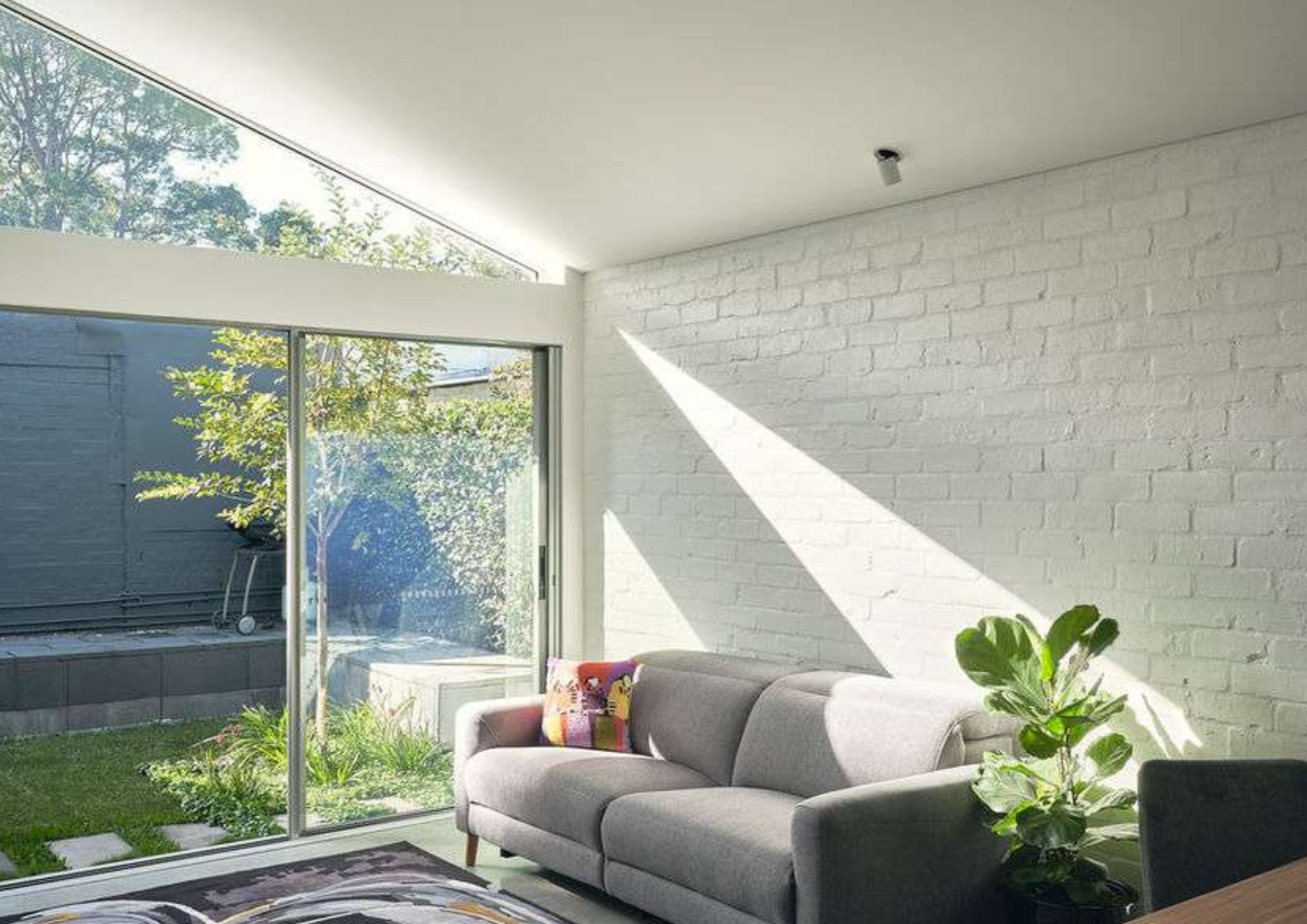 Light-filled living room with a grey sofa, white painted brick wall and large sliding glass doors opening onto a small landscaped courtyard.