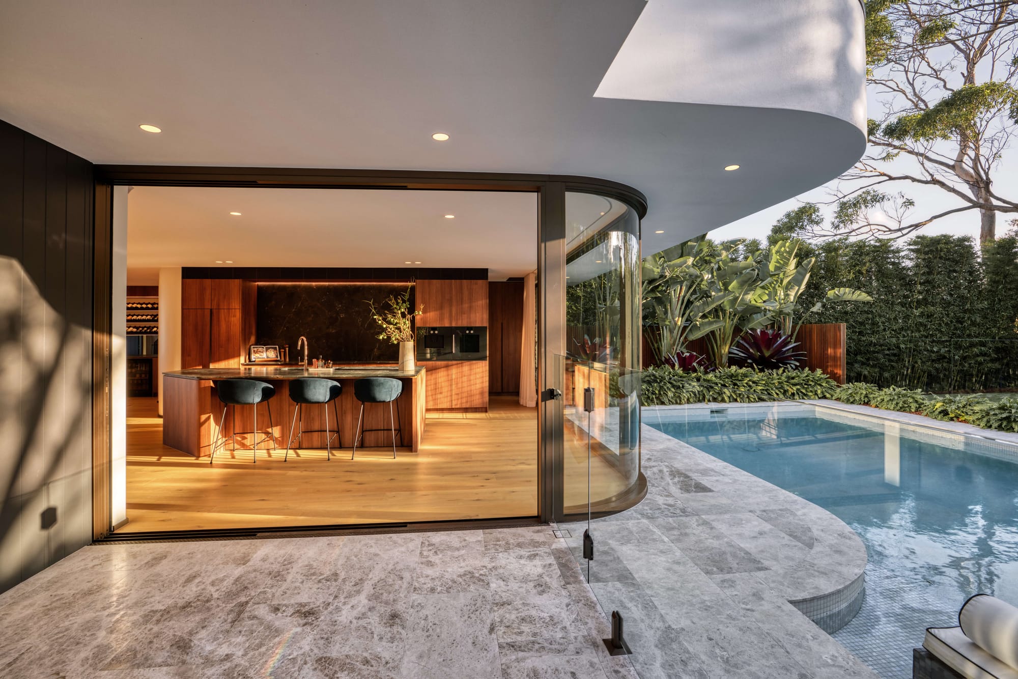 A curved glass door opens from a walnut kitchen with a stone island and black bar stools onto a travertine pool terrace framed by lush tropical planting.