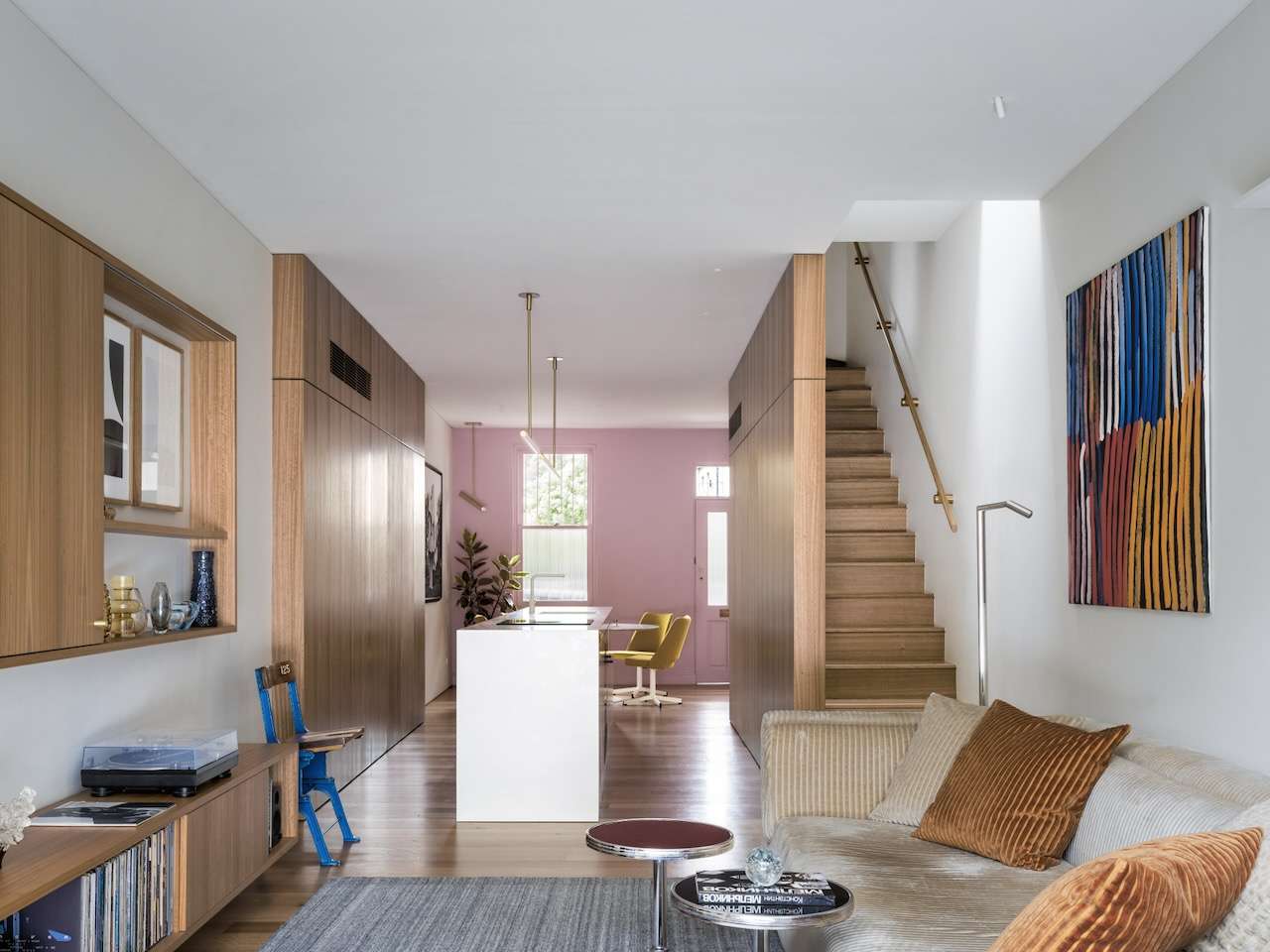 Light-filled living area with timber joinery, pale pink walls, a white kitchen island, timber staircase, neutral sofa and colourful artwork.