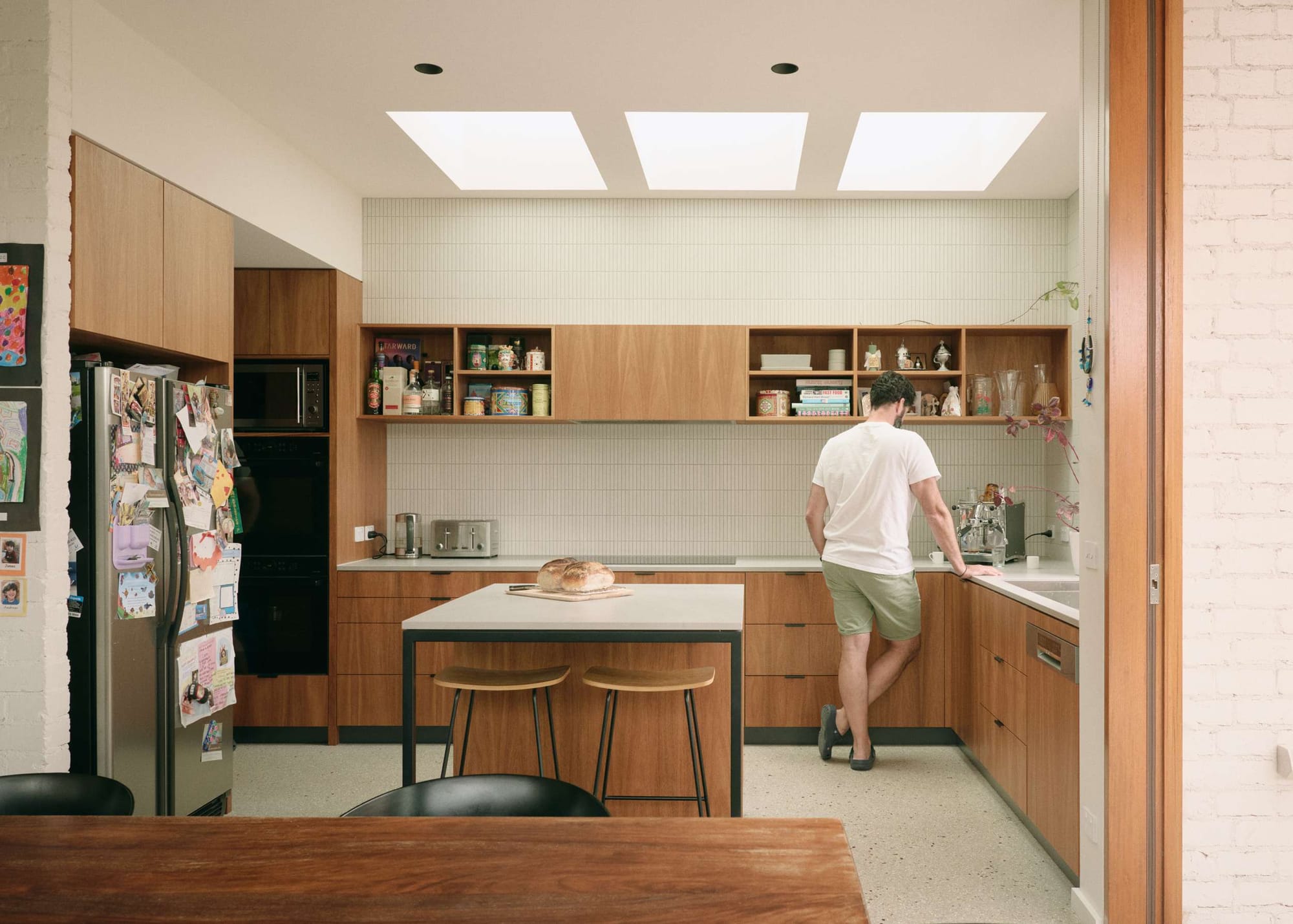 A galley kitchen lit by three ceiling skylights, with walnut cabinetry, open shelving, and a freestanding island; a man stands at the counter near an espresso machine beside a fresh loaf of bread.