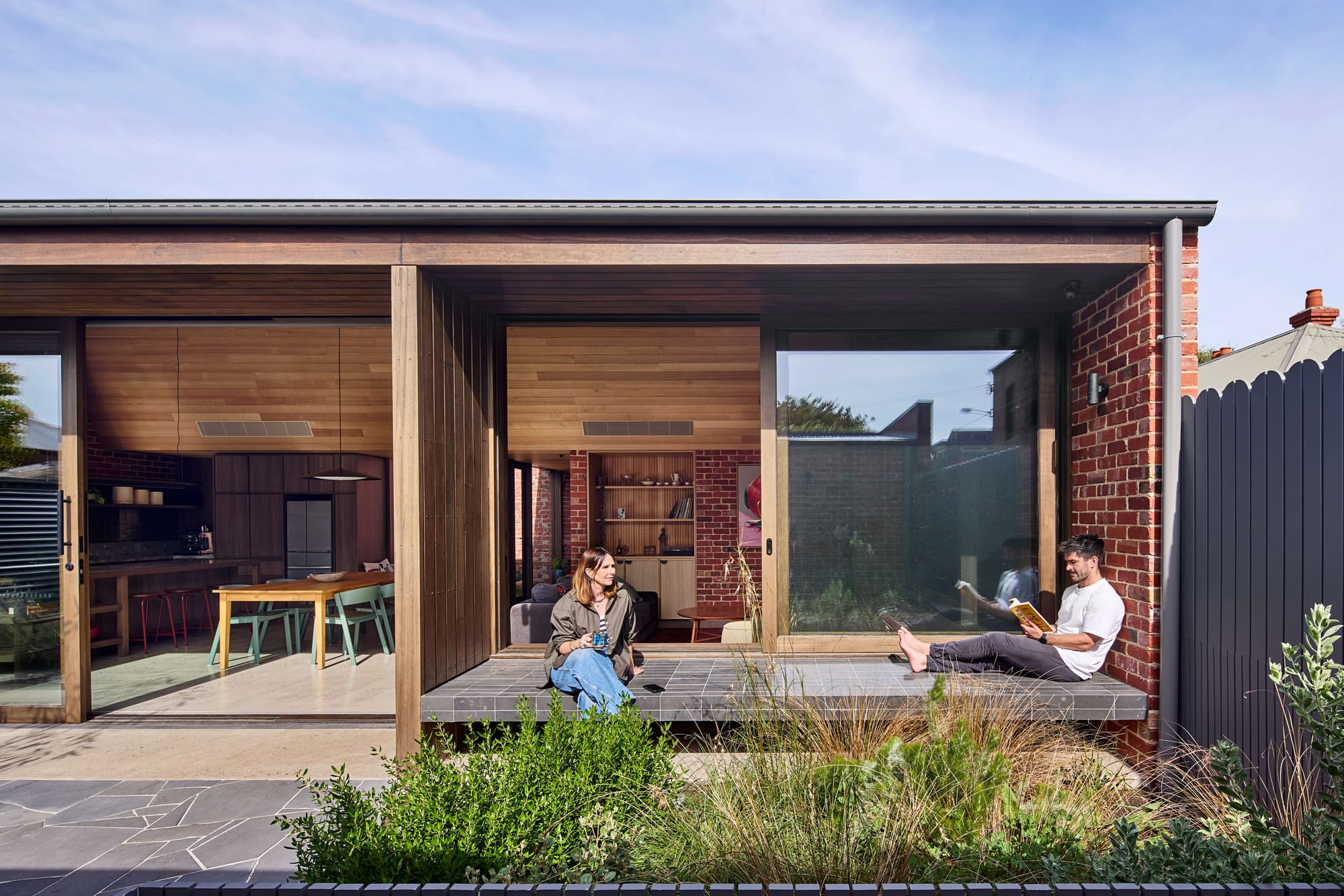 Brick and timber home extension with sliding glass doors open to a tiled outdoor bench, where a woman with coffee and a man reading relax, kitchen and living room visible behind.
