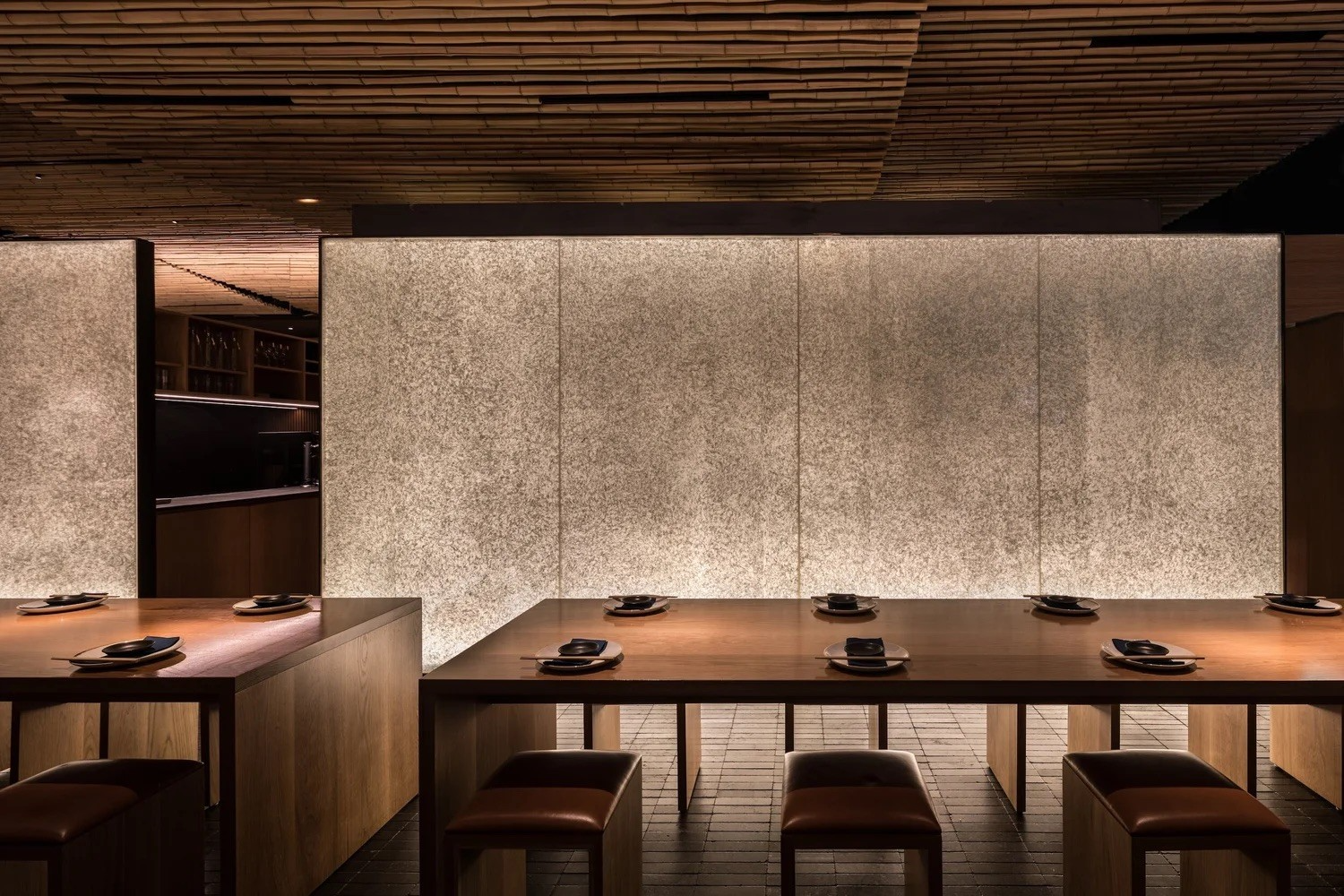 Warmly lit dining interior with timber tables, dark stools, textured illuminated wall panels and a timber-lined ceiling.