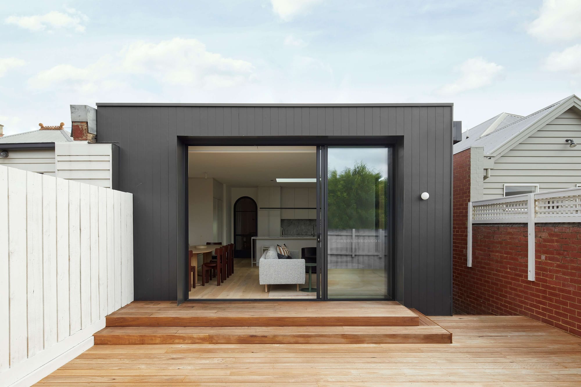 Black-clad rear extension at Richmond Hill House by By Mysa, opening through large sliding glass doors onto a timber deck.