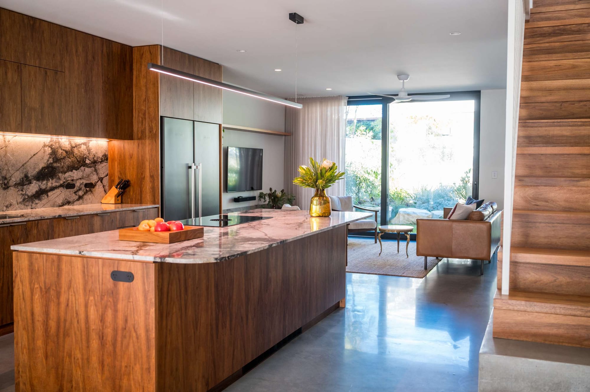 Open-plan kitchen and living area with dark timber cabinetry, a stone island, polished concrete flooring and full-height glazing to the garden.
