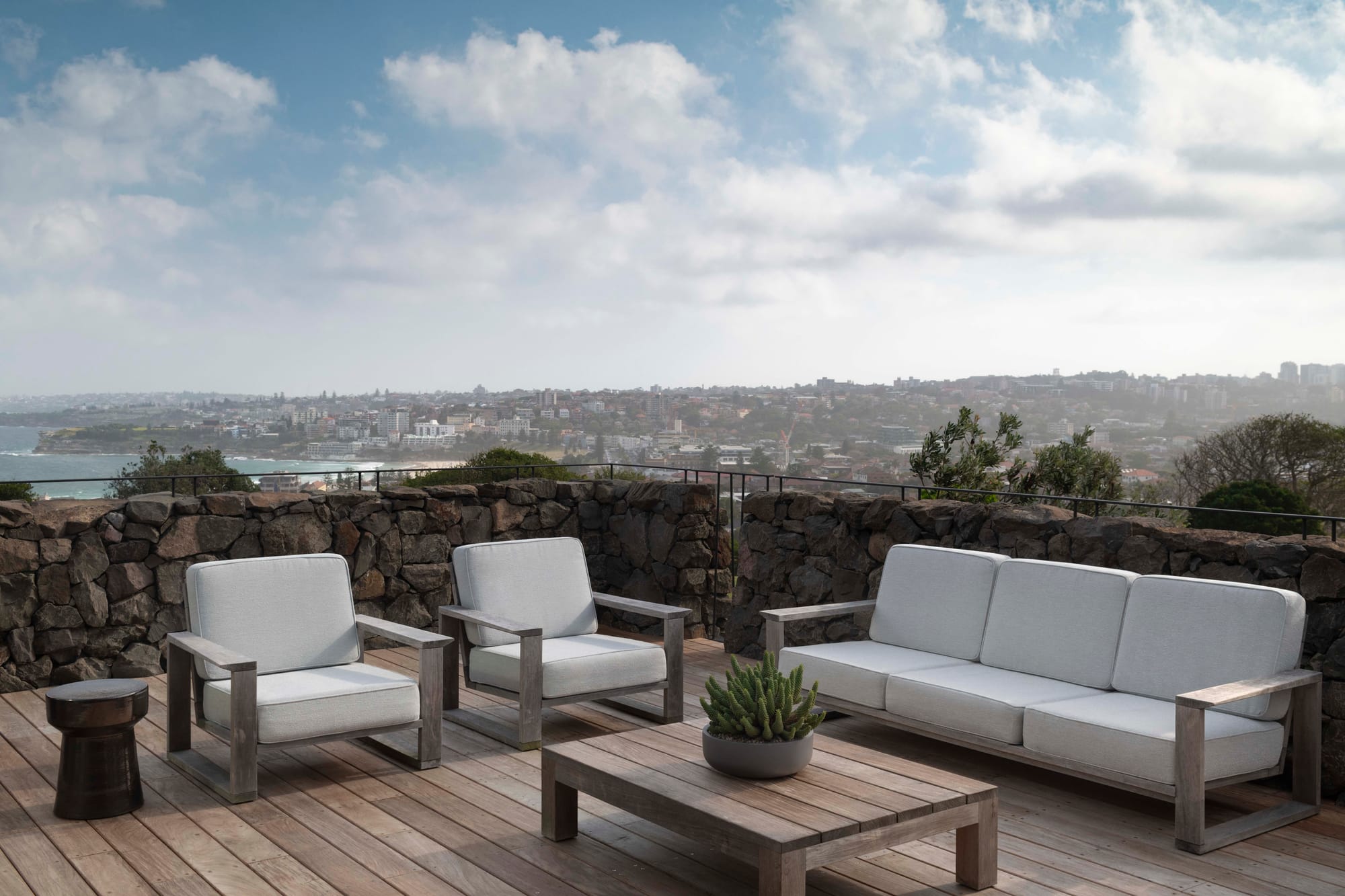 Outdoor lounge setting with timber armchairs and a sofa in pale cushions, a stone retaining wall, and sweeping coastal views over Bondi Beach.