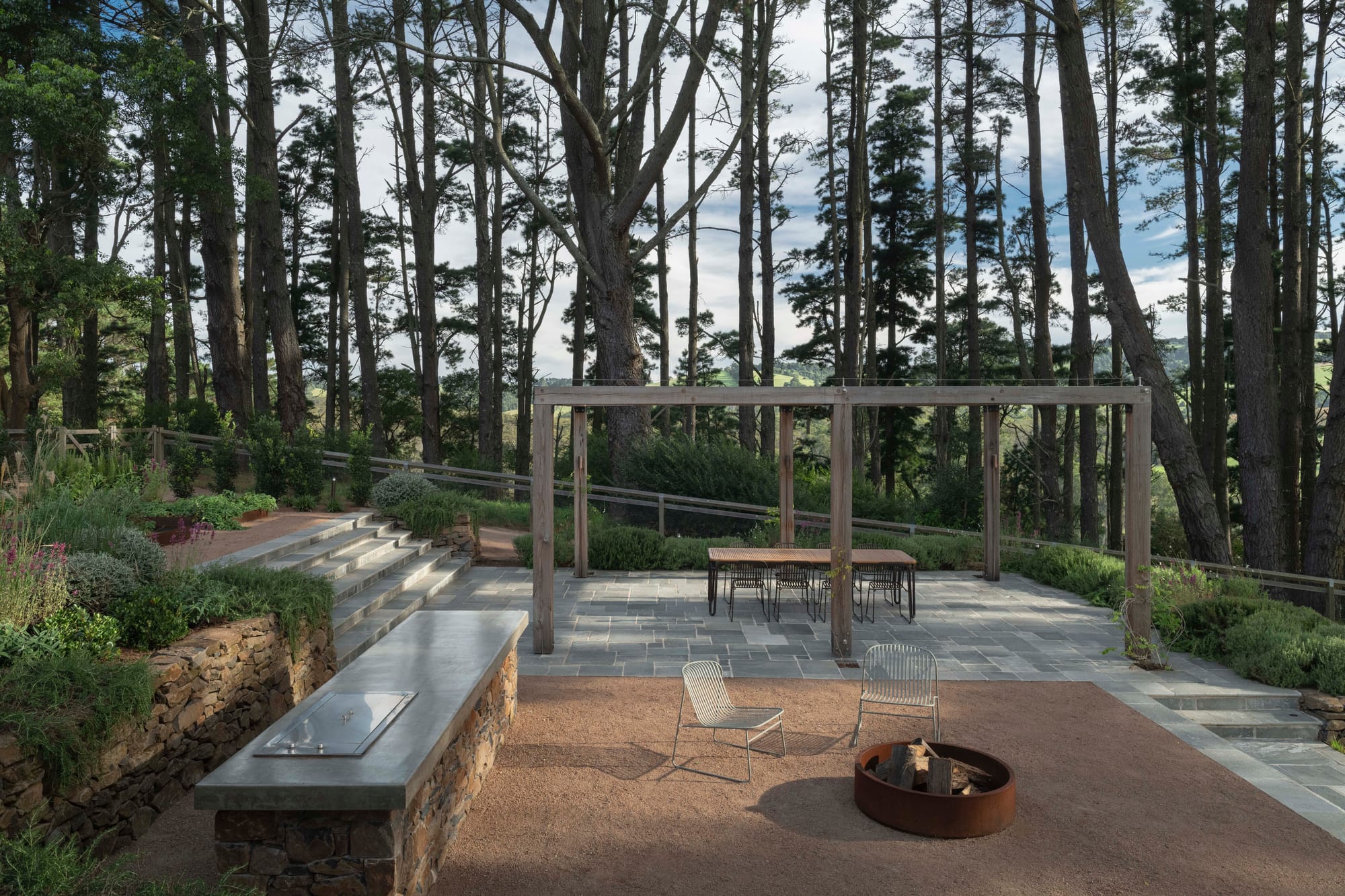 Elevated timber deck with a stone bench, fire pit, outdoor dining table under a timber pergola, surrounded by tall pine trees and rolling hills.