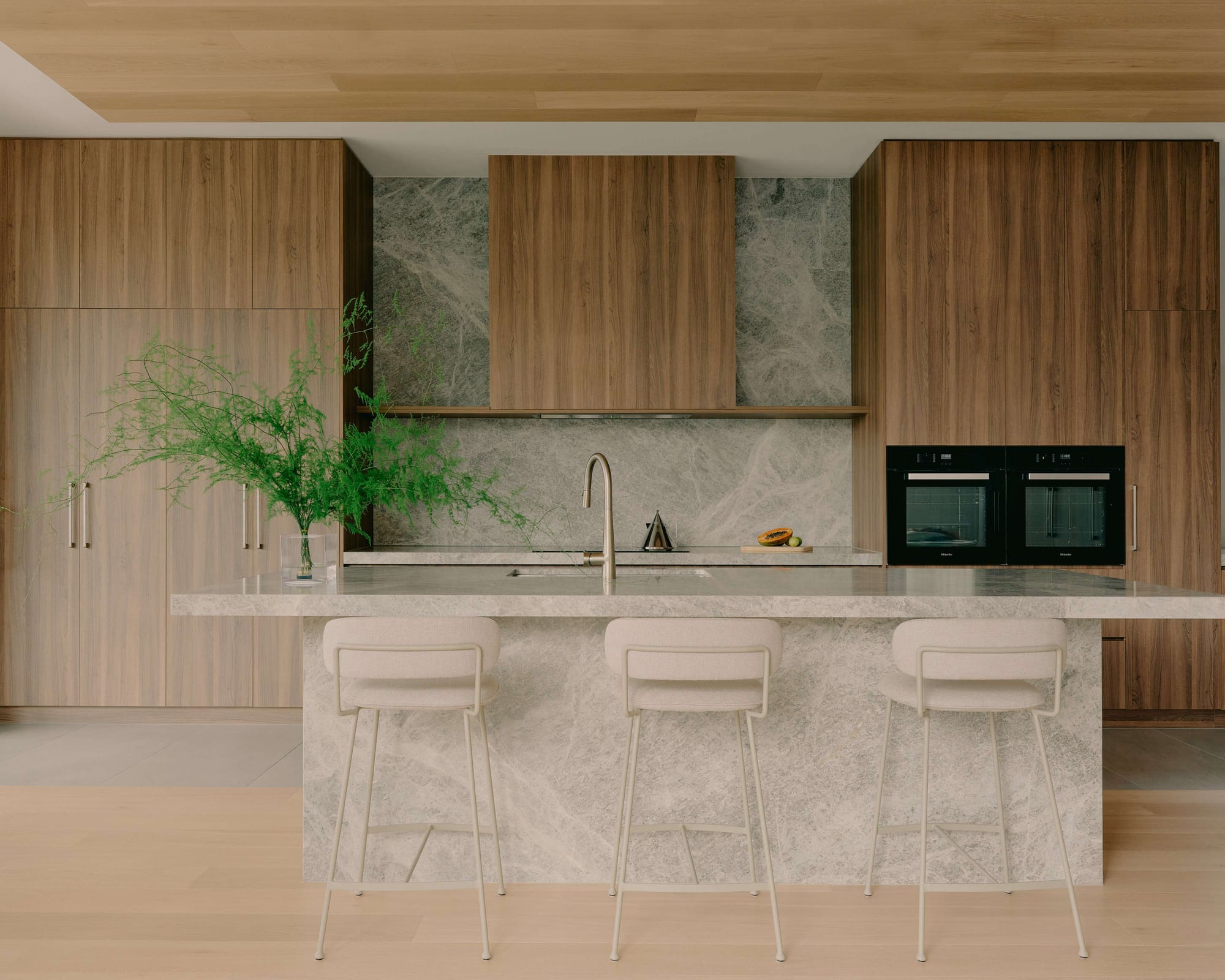 Minimal kitchen with timber cabinetry, stone splashback and island bench, integrated black ovens and three upholstered stools.