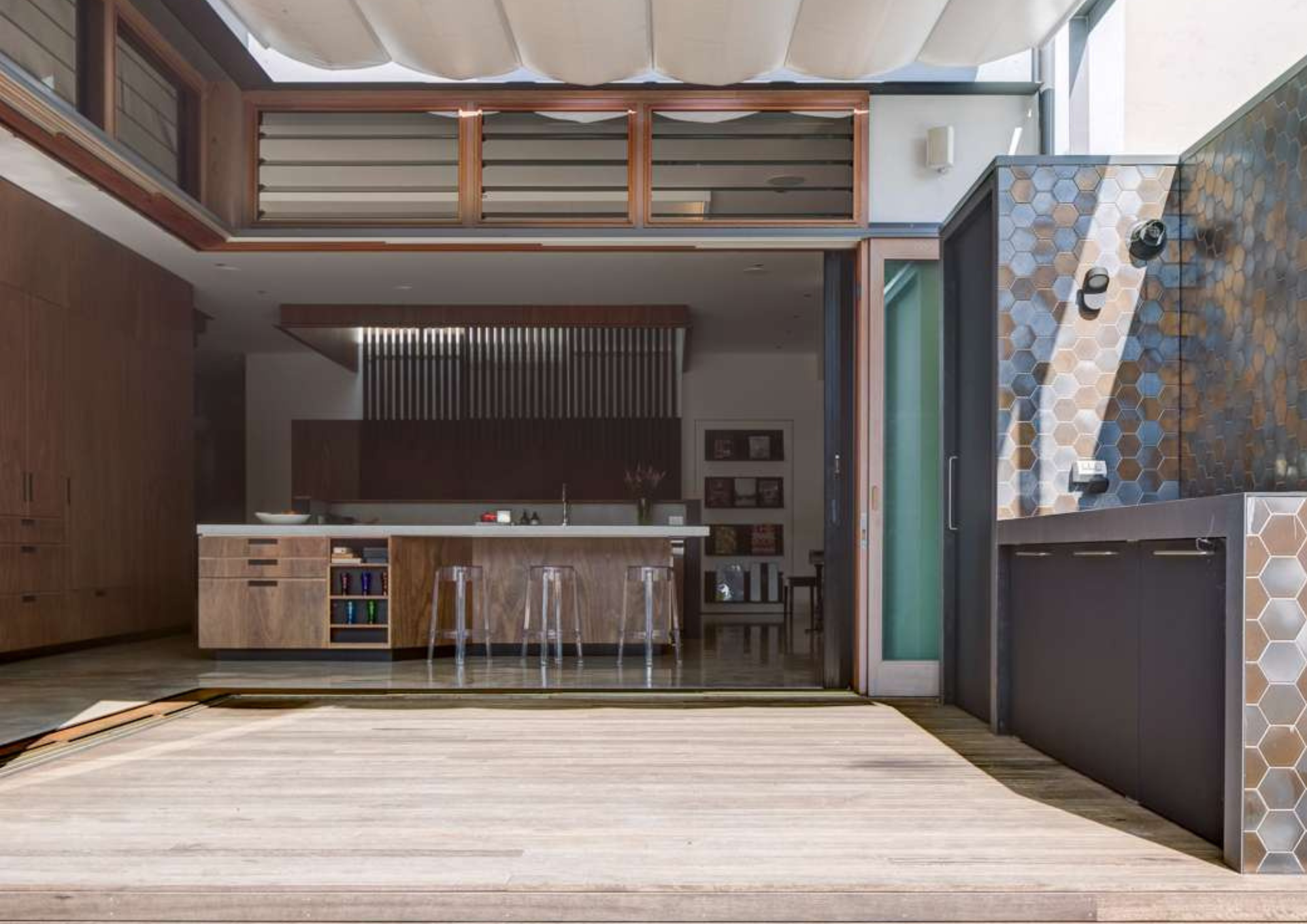 Indoor-outdoor kitchen and entertaining area with timber cabinetry, a stone island bench, retractable canopy and hexagonal tiled outdoor wall.