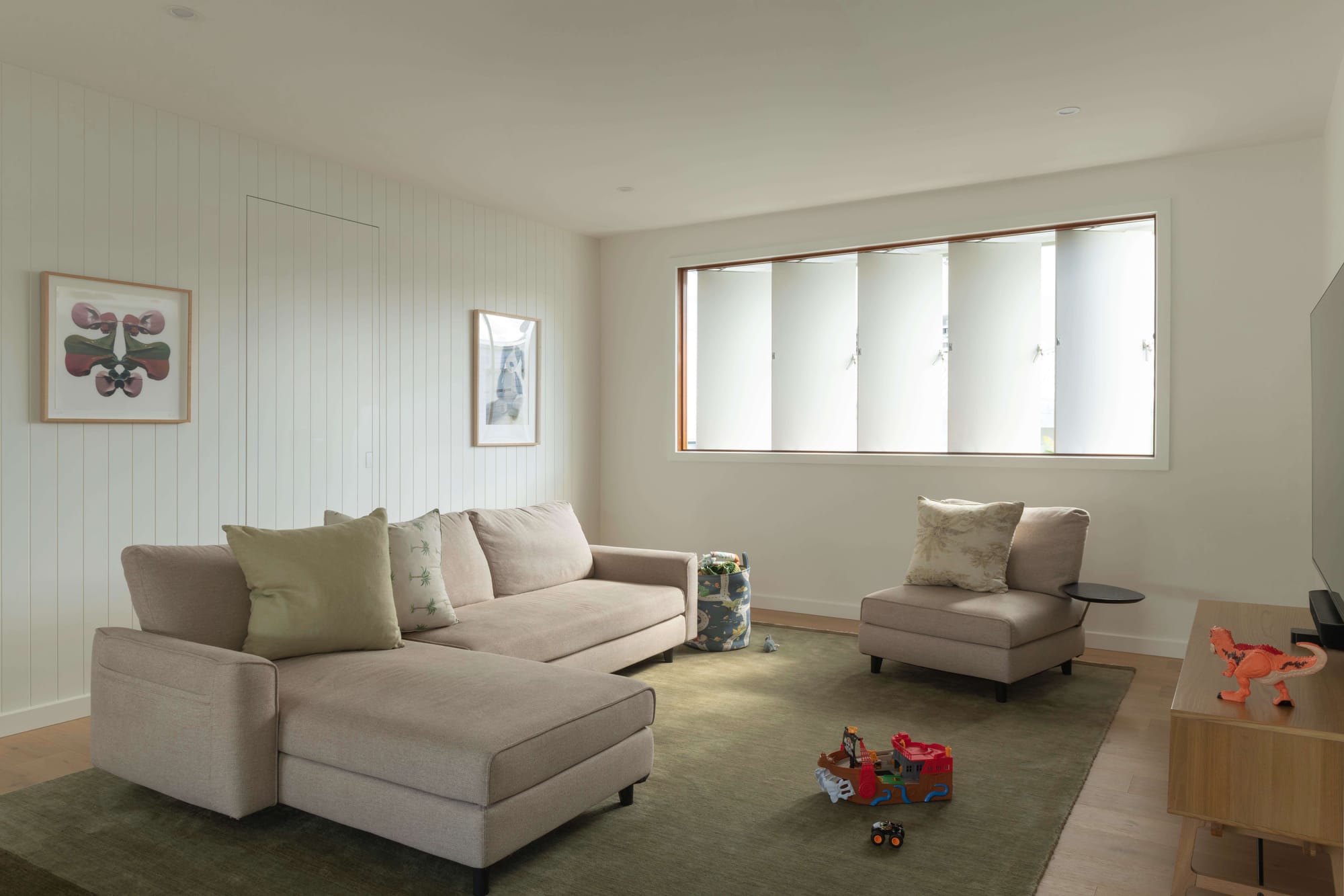 A children's living area with a beige L-shaped sectional on an olive green rug, scattered toys, and white vertical timber-panelled walls hung with framed prints.