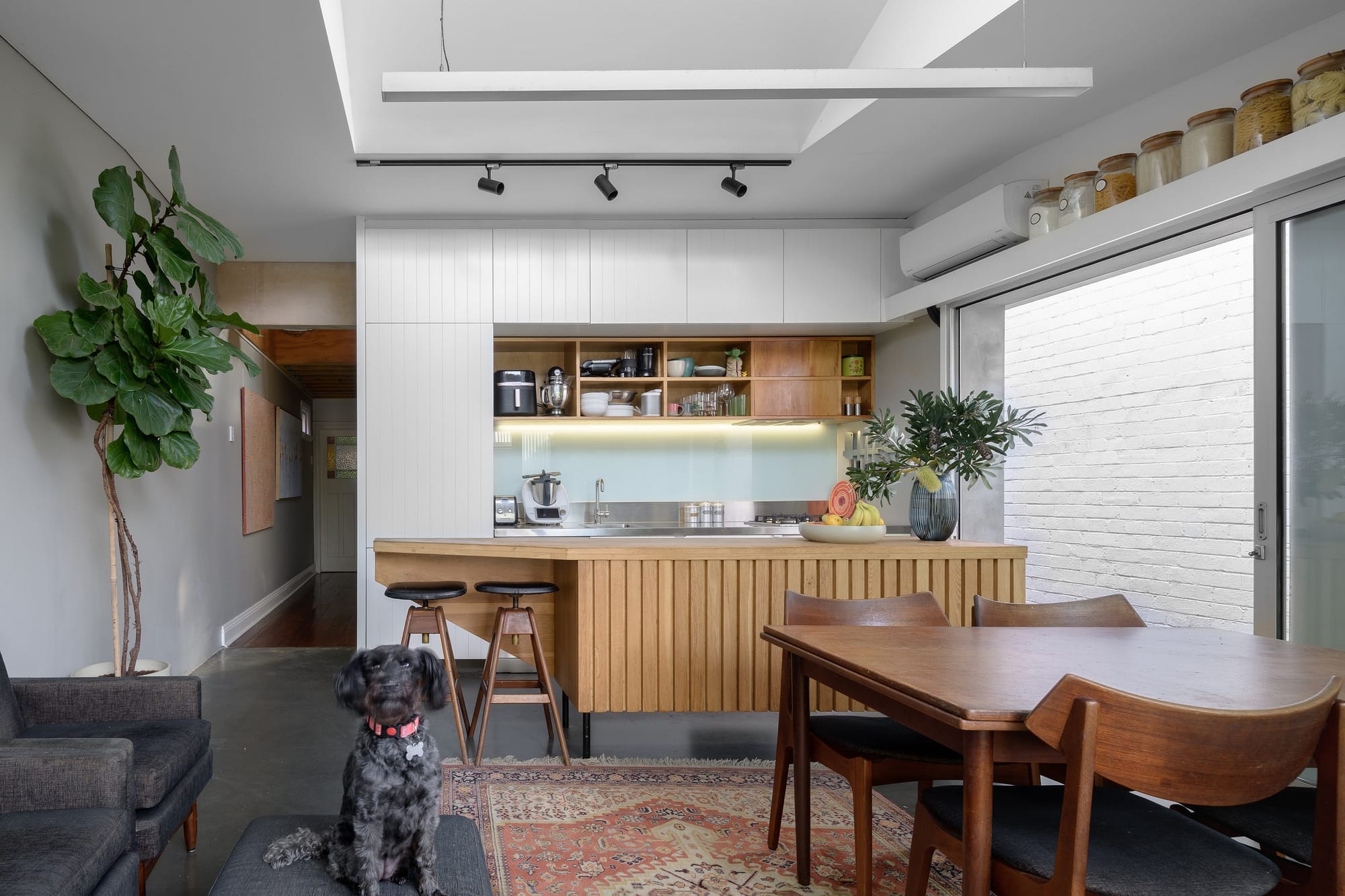Light-filled kitchen and dining area with white cabinetry, timber detailing, open shelving, a slatted timber island and a black dog seated nearby.