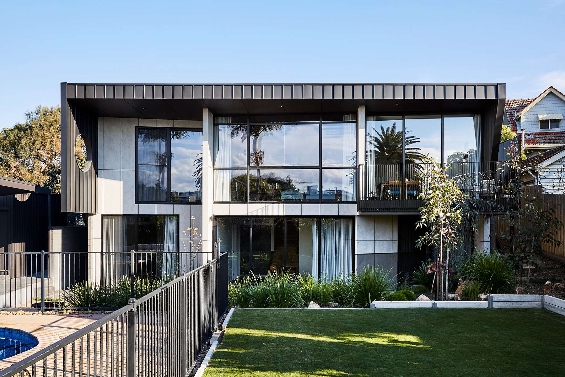 Two storey contemporary home with dark cladding, floor to ceiling glass windows, a balcony and a landscaped garden with a swimming pool in the foreground.