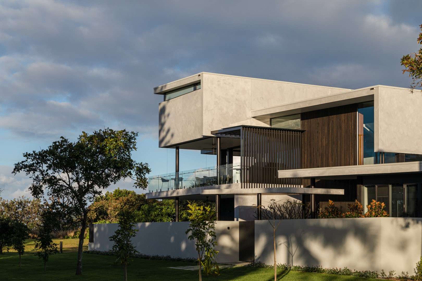 A large contemporary home with rendered upper-level cantilevers, timber louvred screens, and a glass-balustraded balcony, set against a cloudy sky and lawn.