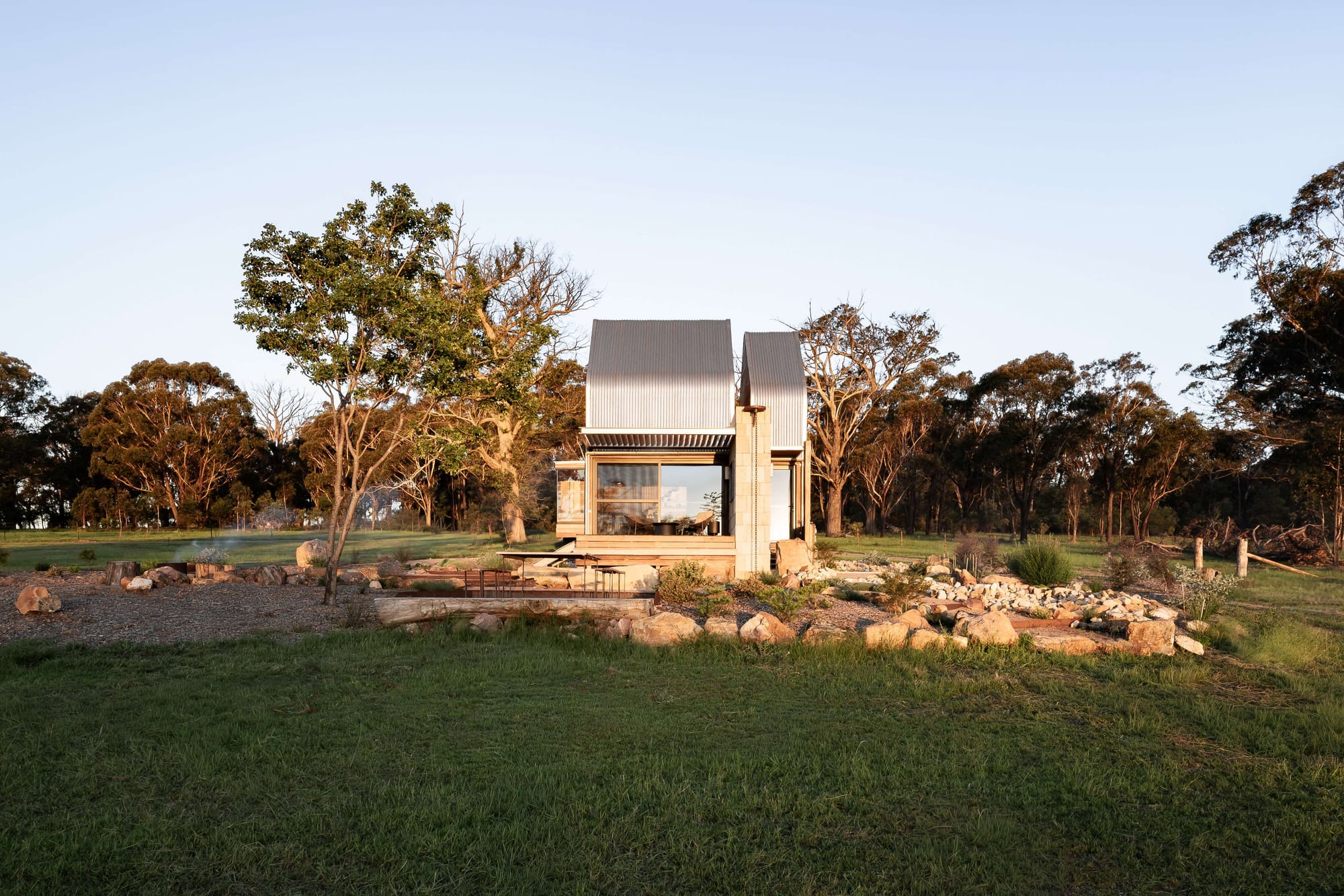 Small corrugated metal cabin by AO Design Studio set among grass, rocks and mature trees in the Australian landscape.