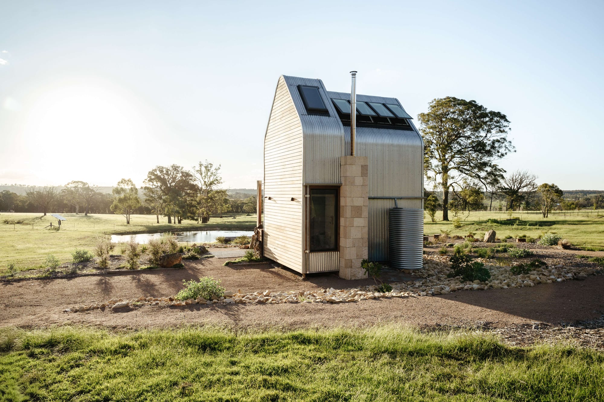Compact corrugated metal cabin by AO Design Studio with a curved roof, rooflights and water tank set within a rural landscape.