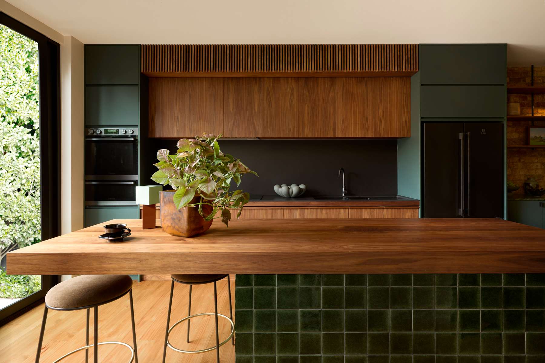 Dark green and timber kitchen with a thick timber island bench, green tiled base, integrated appliances and a large indoor plant.