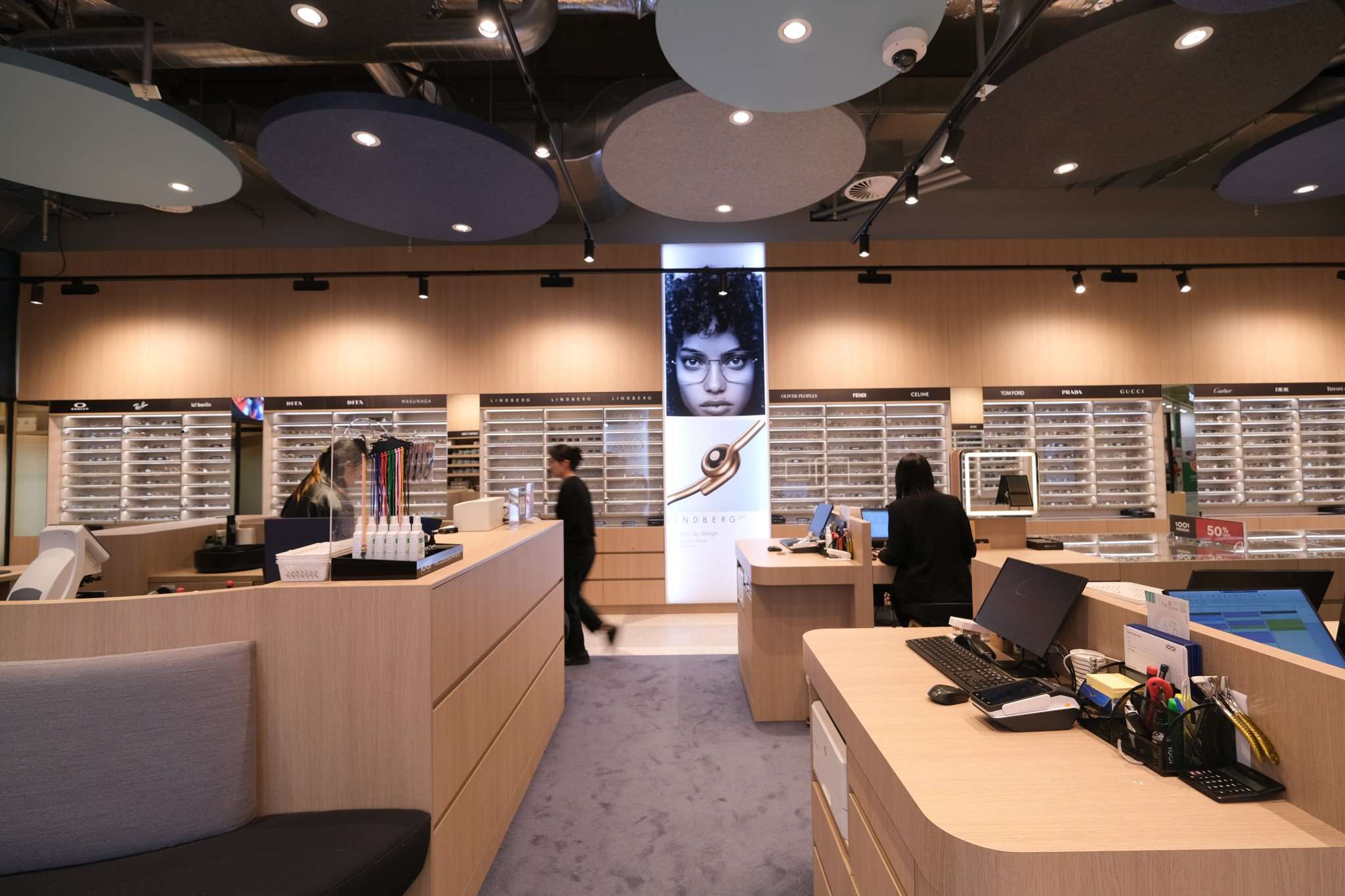 Optical store interior with pale timber counters, illuminated eyewear displays, circular acoustic ceiling panels and staff assisting customers.