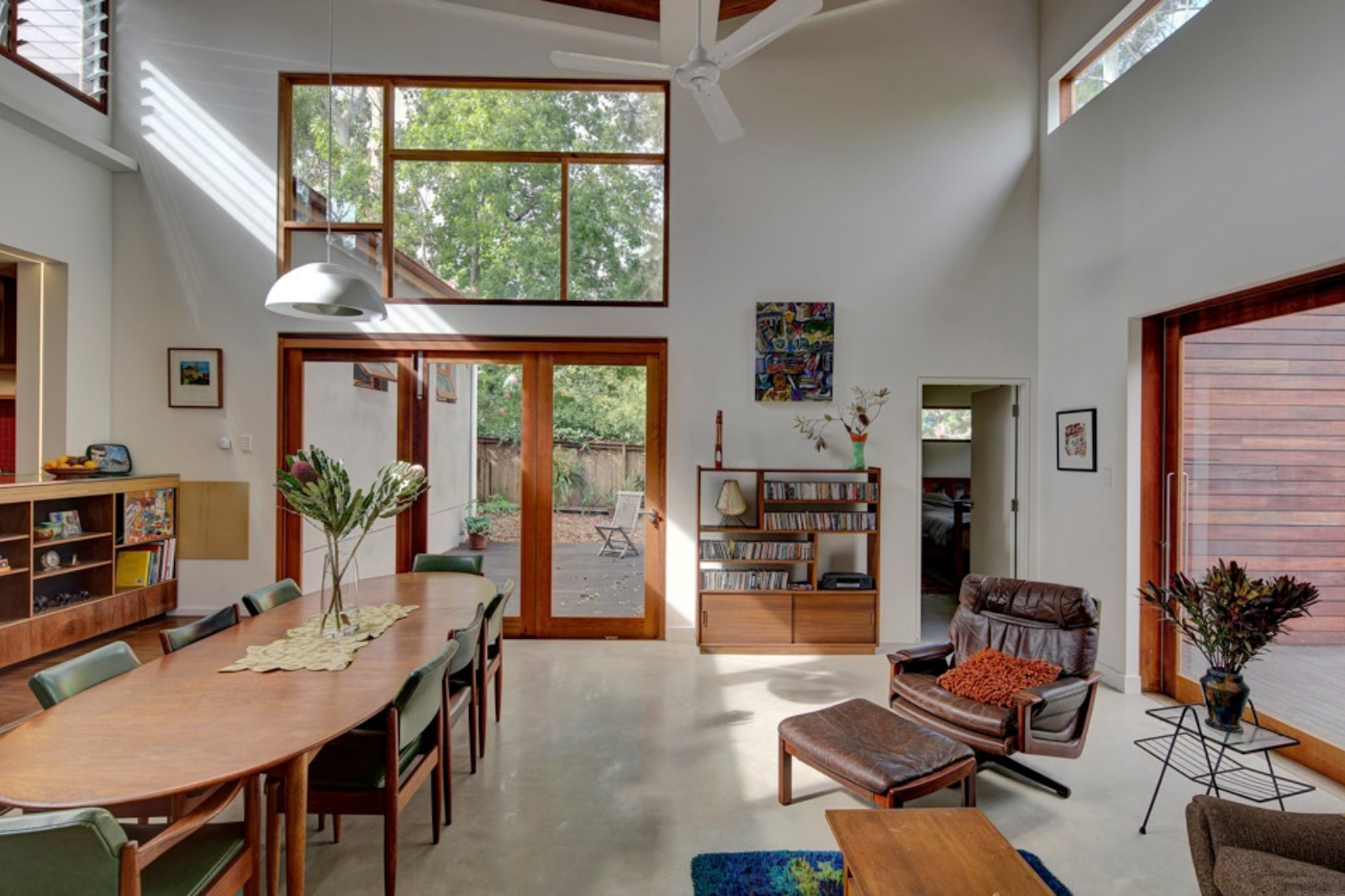 Light-filled living and dining room at Breuer House, with high windows, timber furniture and glazed doors opening to a courtyard.