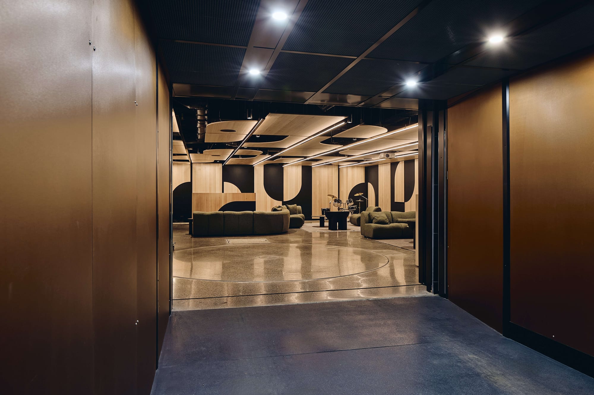 Lounge and music space with green sofas, a drum kit, geometric timber wall panels and polished concrete flooring.