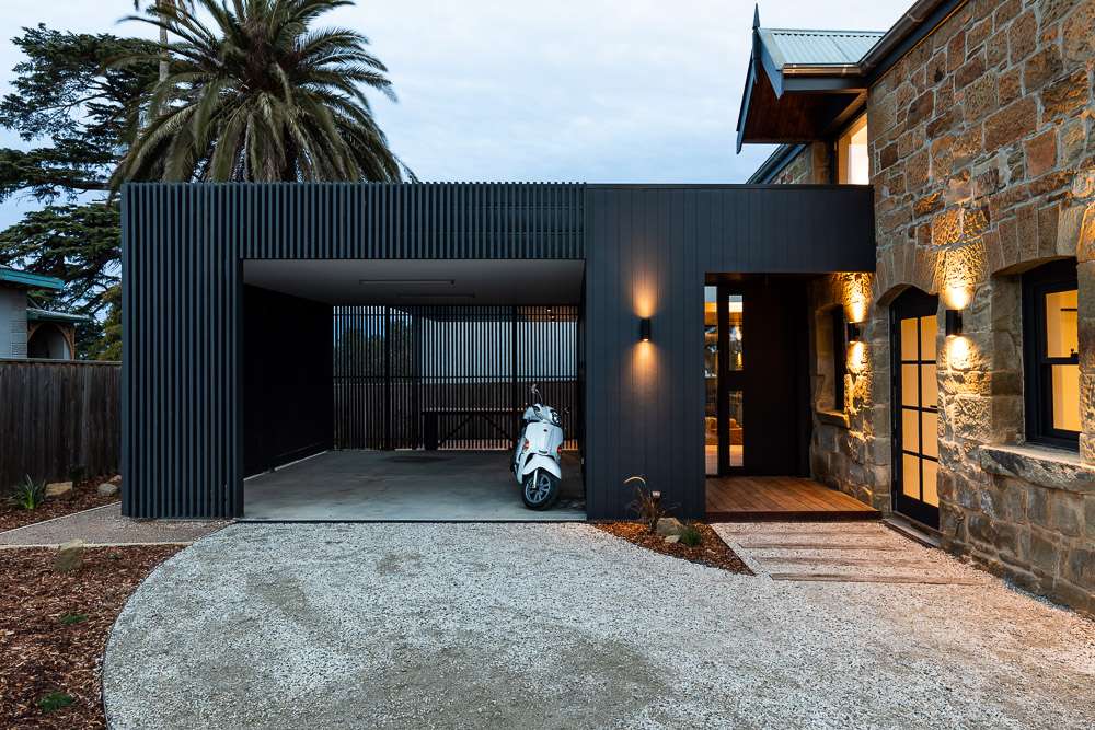 Contemporary black carport adjoining a stone house, with a white scooter, timber entrance deck and curved driveway.