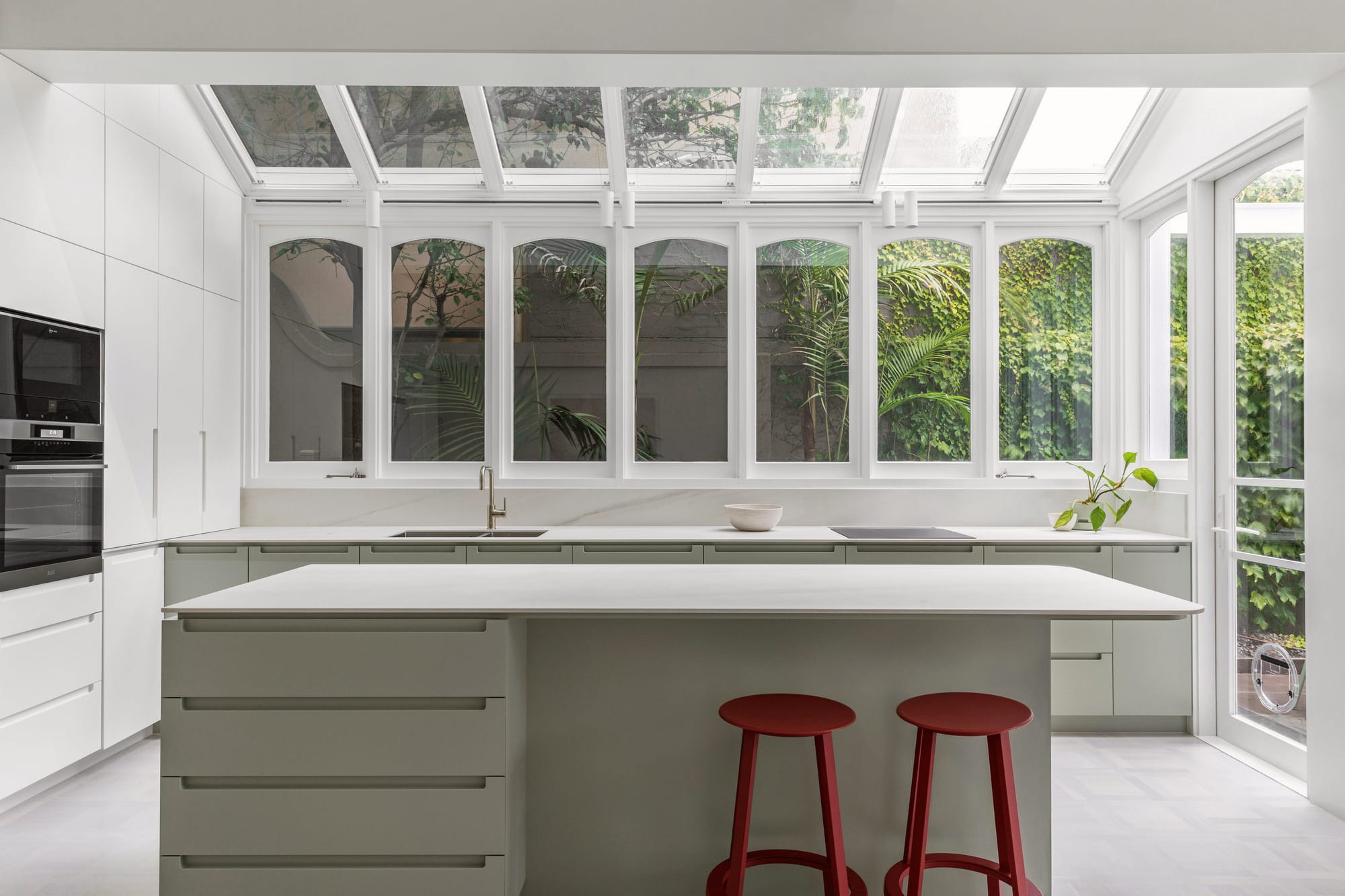 Light-filled kitchen at Canterbury Road House by Ben & Ben with pale green cabinetry, a large island and glazed walls and roof overlooking the garden.