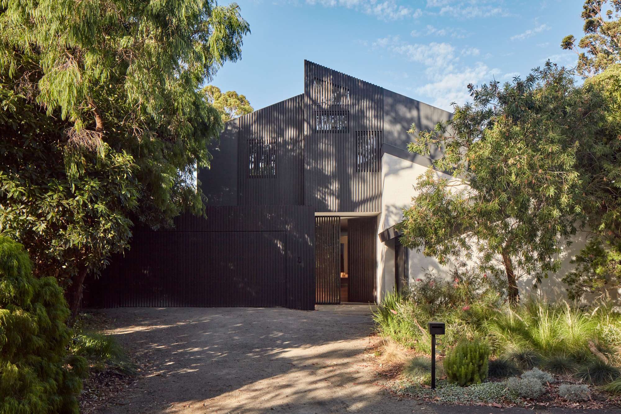 Contemporary home with dark vertical facade screening, an angular roofline and a recessed entrance surrounded by mature trees.