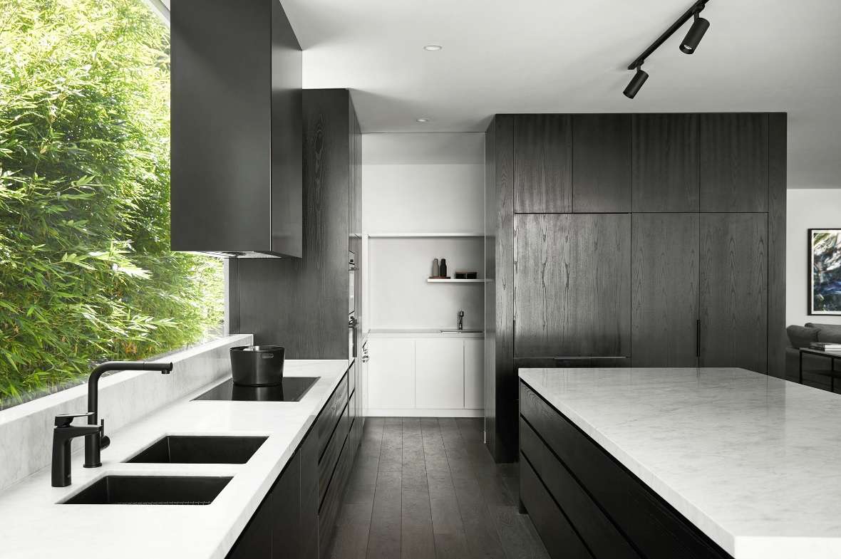 Black timber kitchen by Joseph Galati Architecture with white benchtops, twin sinks and a long window framing dense green planting.