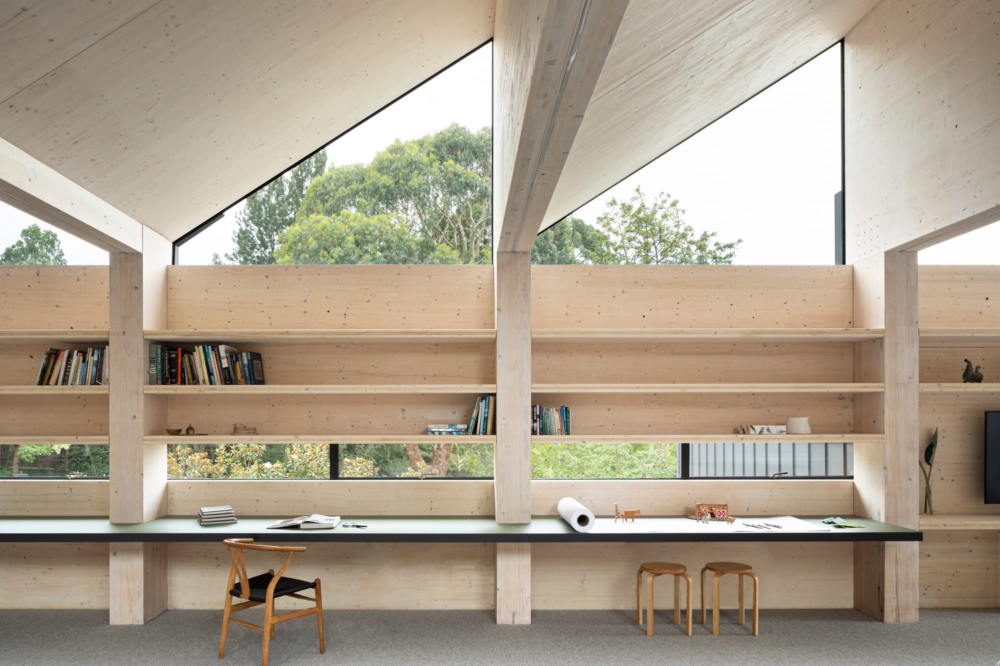 Timber-lined study with exposed beams, triangular clerestory windows and built-in bookshelves above a long green desk.