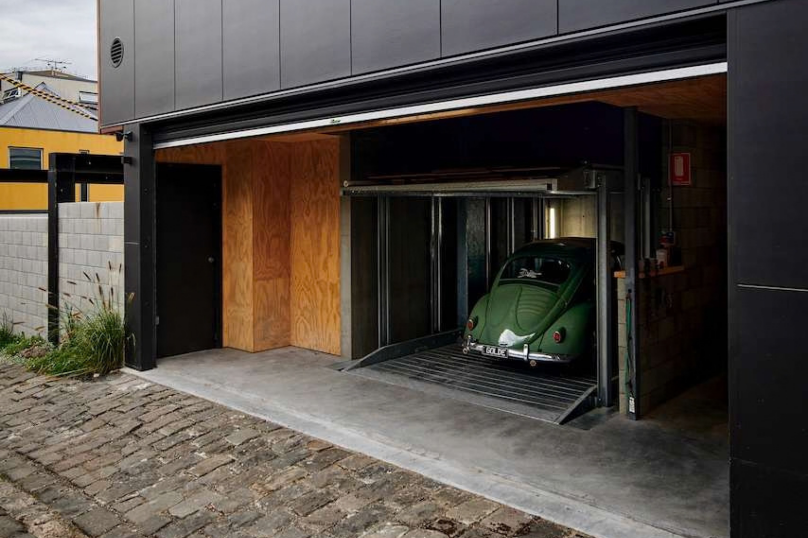 Green vintage Volkswagen Beetle on a vehicle lift inside a garage with plywood wall lining and dark exterior cladding.