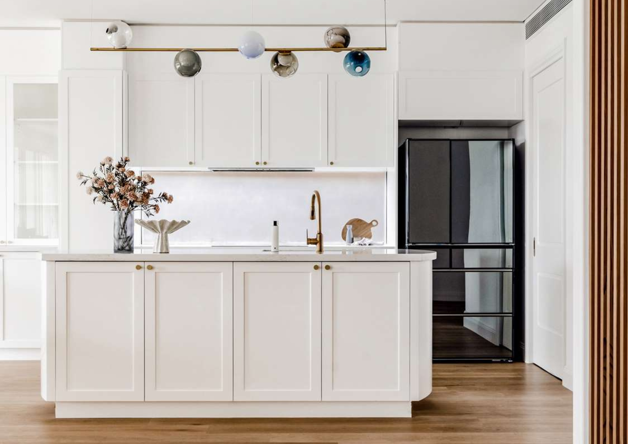 Bright kitchen with white panelled cabinetry, a curved island end, brass tapware and timber flooring at Clanwilliam House by Milli Studio.