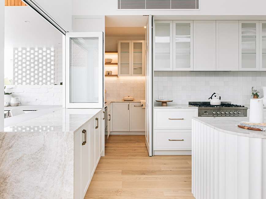 Bright white kitchen by Blank Creatives with stone benchtops, a fluted island and an open doorway leading to a concealed pantry.