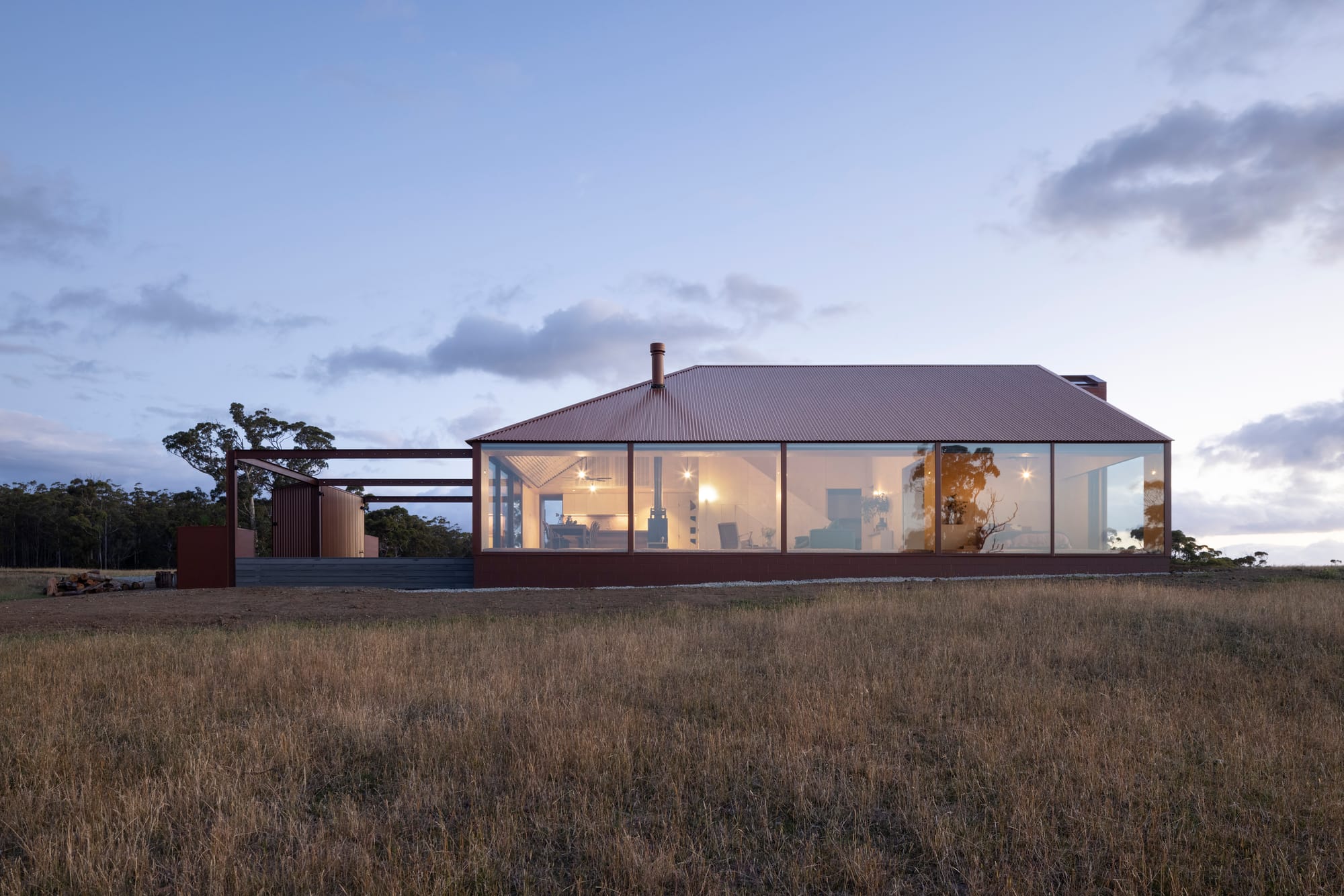 Rural home at dusk with a red corrugated roof, full-height glazing and an adjoining pergola overlooking open grassland.