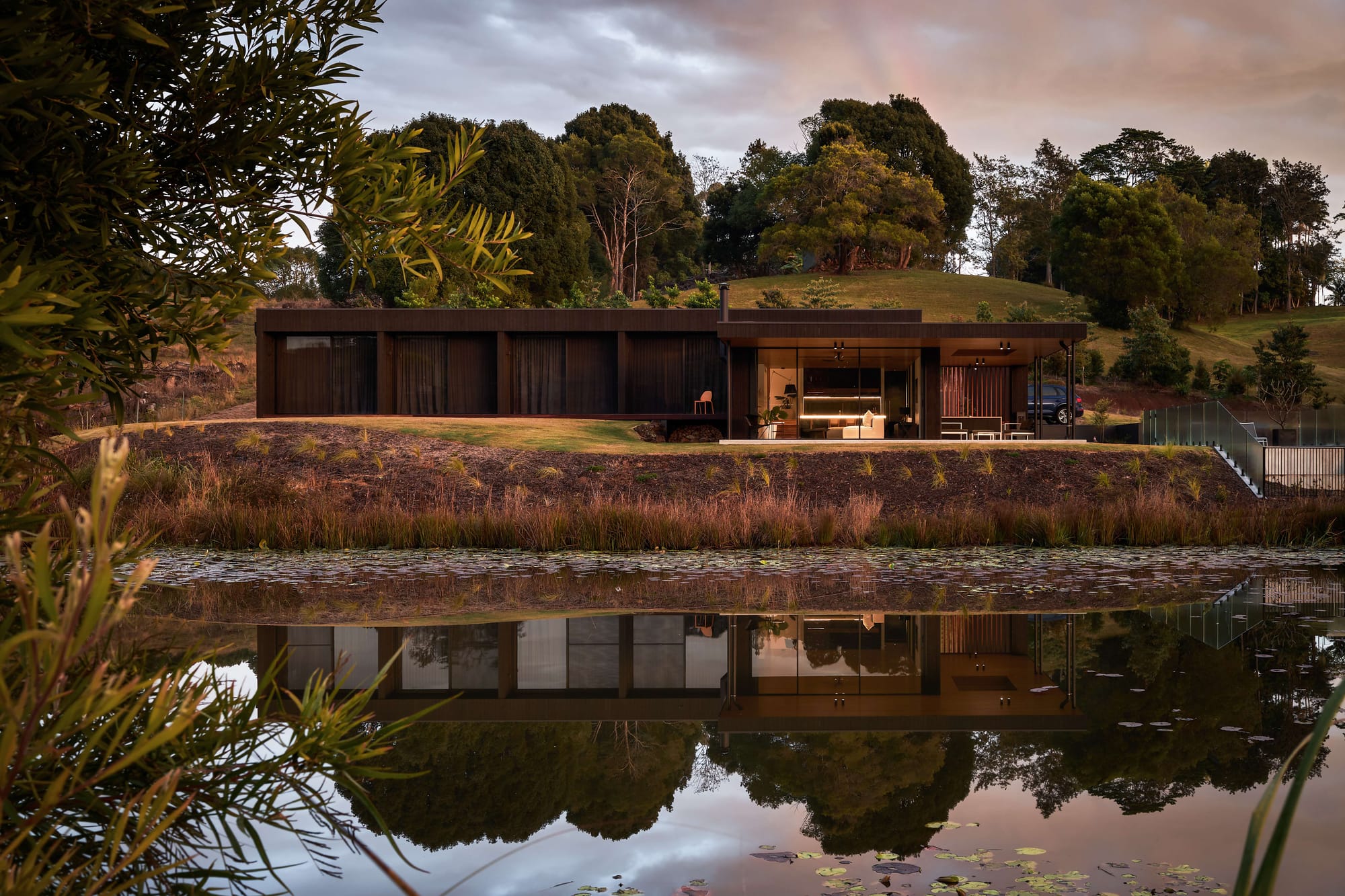Low dark-clad home overlooking a reflective pond and landscaped hillside at Coorabell Springs. Photography by Andy MacPherson.