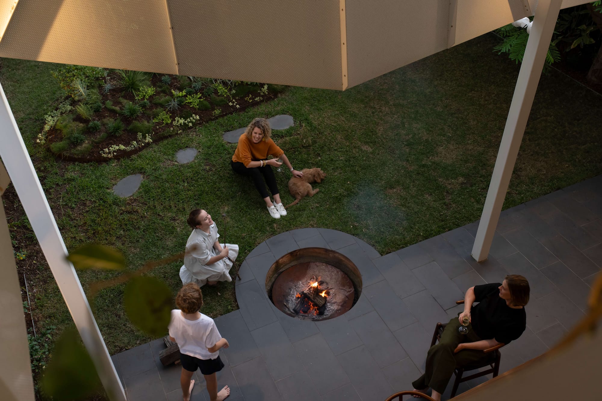 Family gathered around a circular outdoor fire pit within the landscaped garden at Cornerstone by LOUPE Architecture. Photography by Alanna Jayne McTiernan.