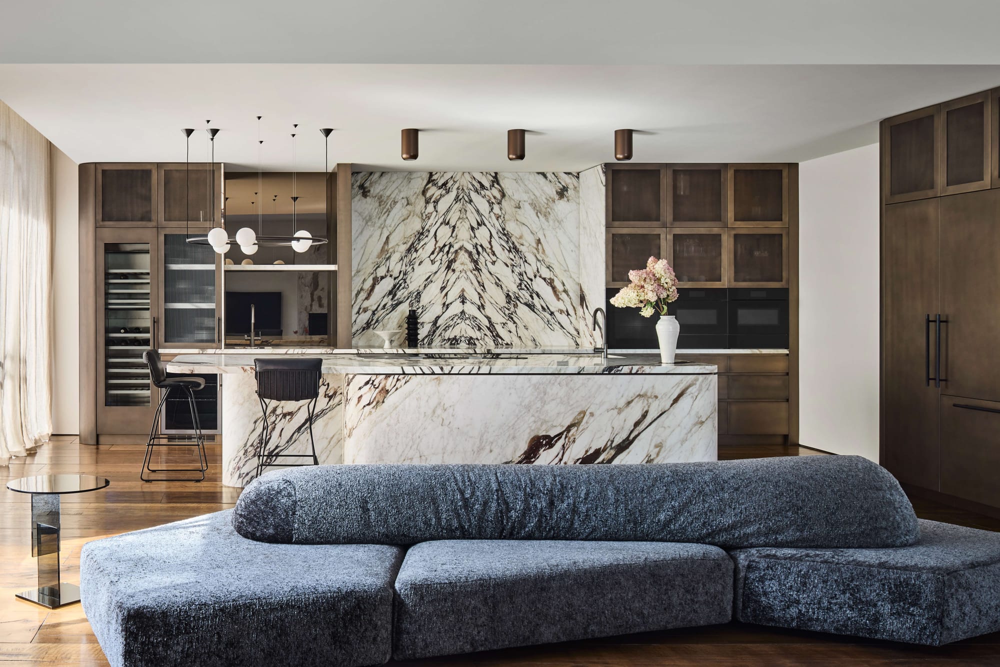 Open plan kitchen and living area at Davis House in Melbourne by AKI Design, featuring bookmatched Viola marble, dark timber cabinetry and a curved blue sofa.