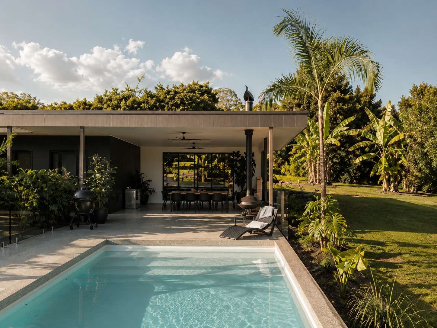 In-ground swimming pool beside a covered outdoor pavilion with a timber ceiling, dining table and tropical planting, by Sarah Waller Architecture.